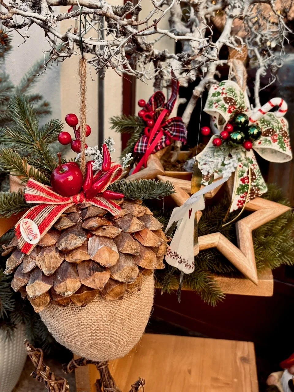 Decorazioni natalizie con coni di pino, fiocchi rossi, bacche rosse, e ornamenti a forma di stella e angelo, su un ramo innevato con pigne e tessuto a sacchetto naturale.