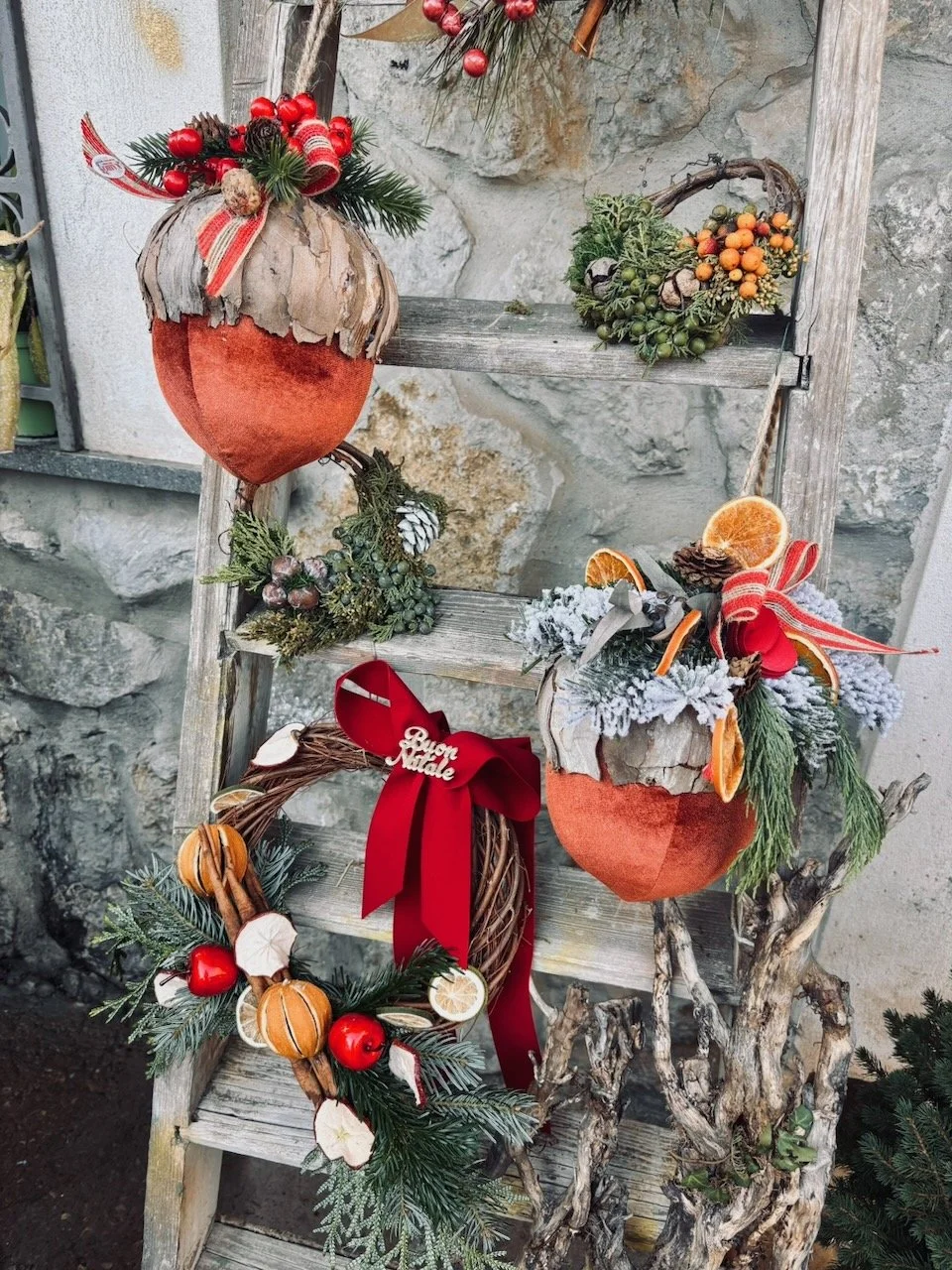 Decorazioni natalizie su una sedia di legno, tra cui ghirlande con frutta, pigne, bacche, rami di pino, e un mazzo con un fiocco rosso con la scritta 'Buon Natale'.