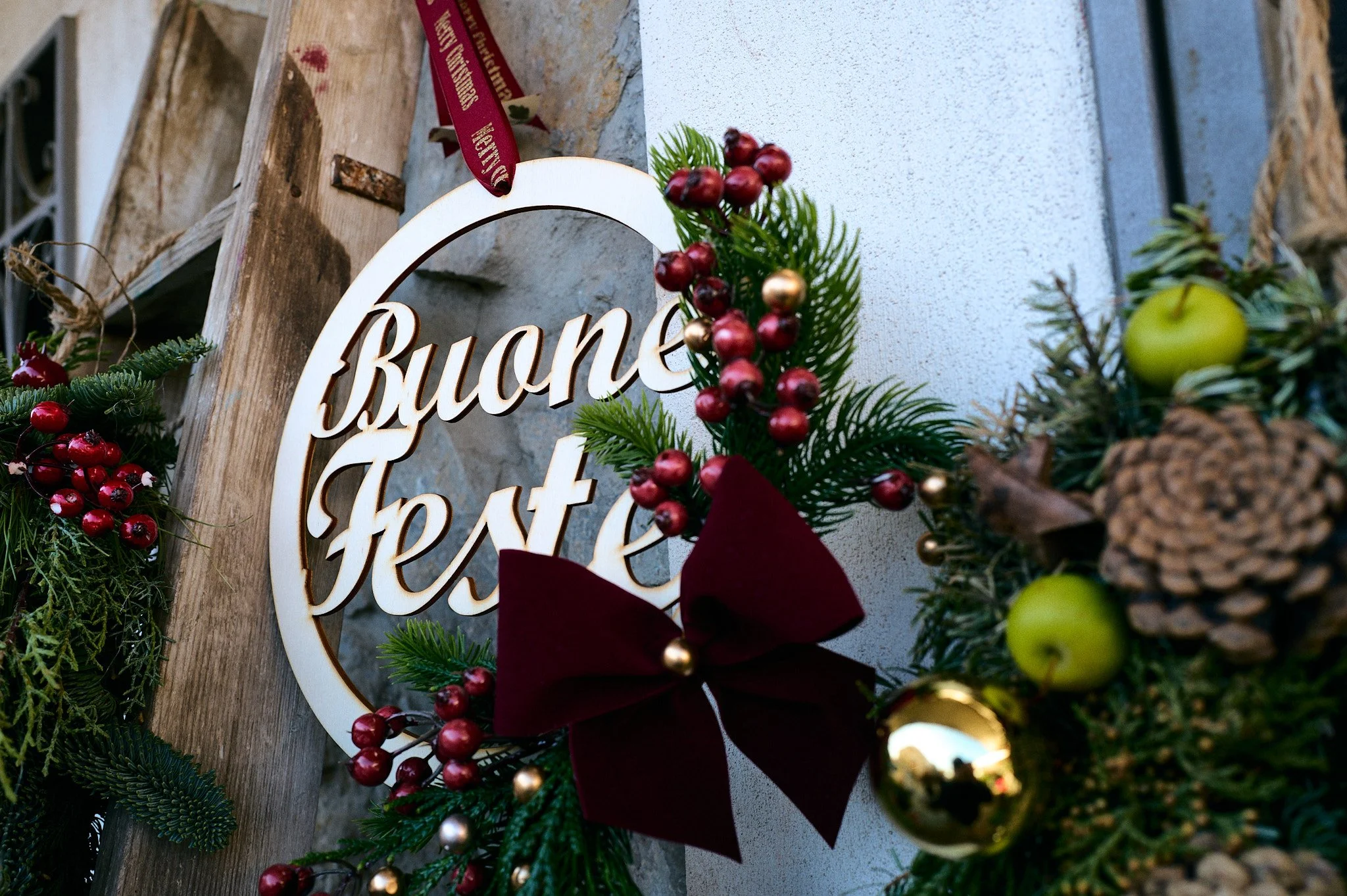 Decorazioni natalizie con un cartello che dice 'Buone Feste', foglie di abete, bacche rosse e dorate, un fiocco di velluto rosso, pigne, mele verdi e una sfera dorata.