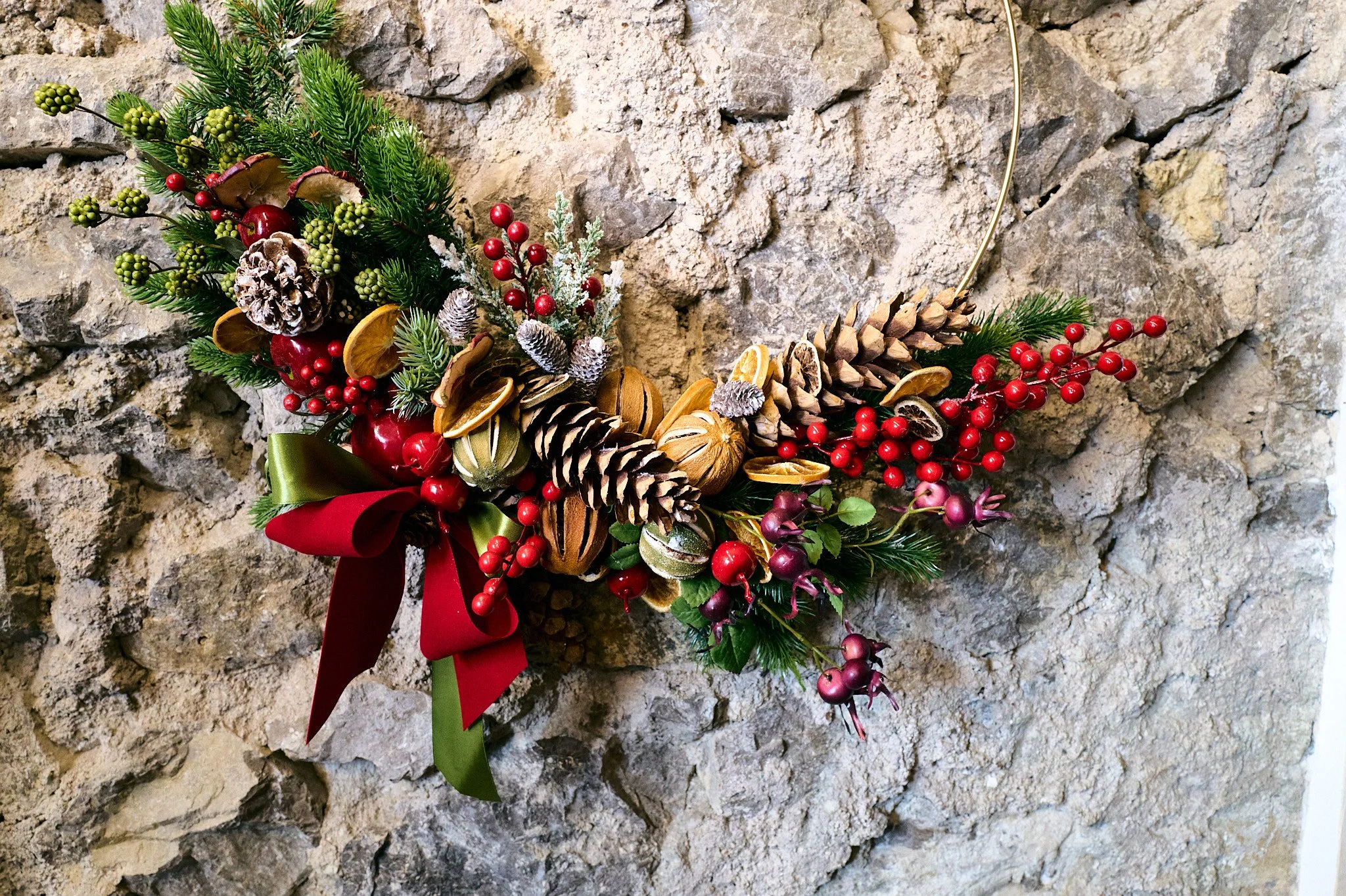 Corona natalizia decorata con pigne, rami di abete, bacche rosse, agrifoglio, e nastri rosso e verde, appesa a un muro di pietra.