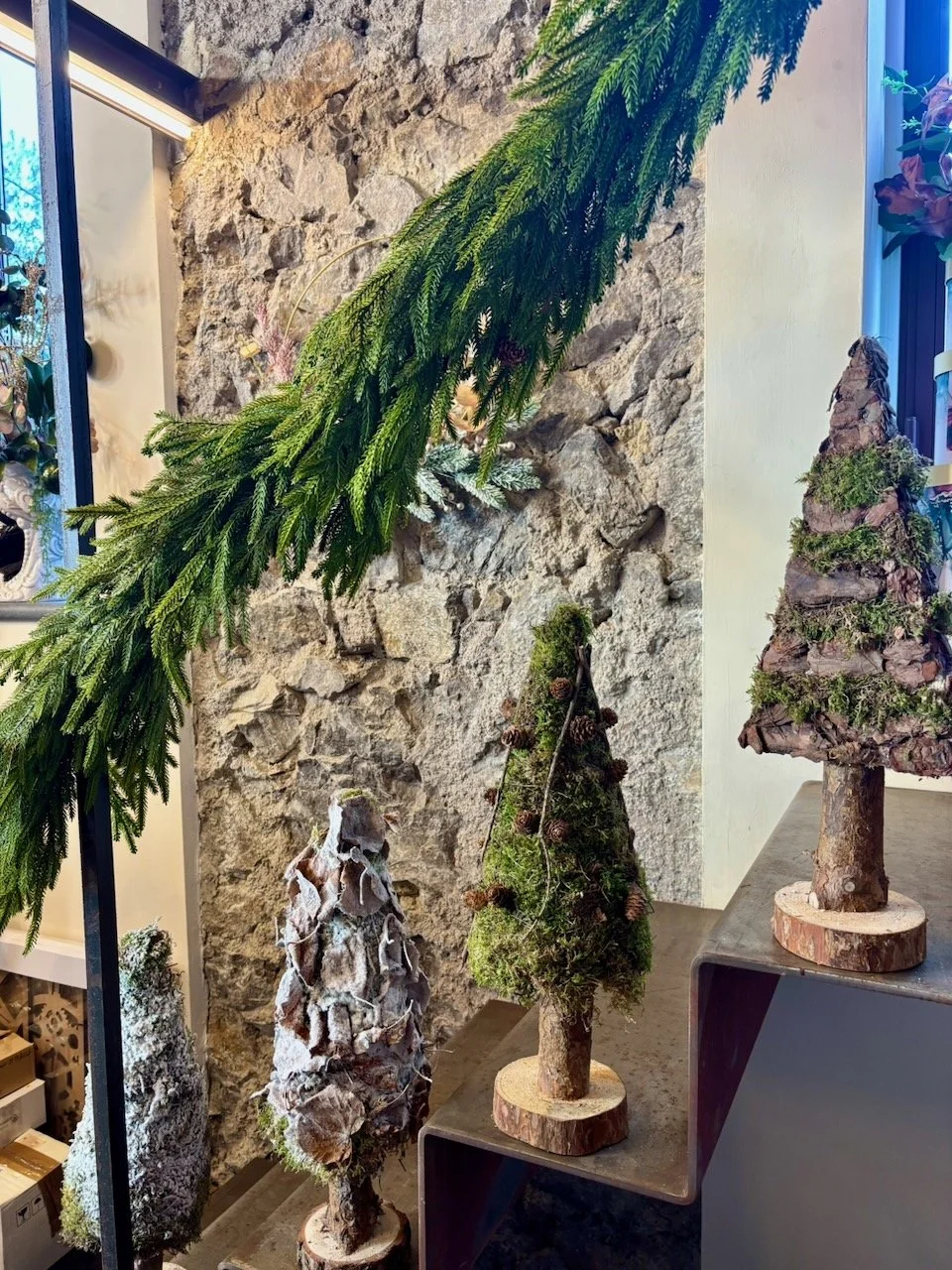 Tre alberelli di Natale realizzati con tronchi e foglie di vari materiali decorativi, posizionati su una scala in un ambiente con parete di mattoni.