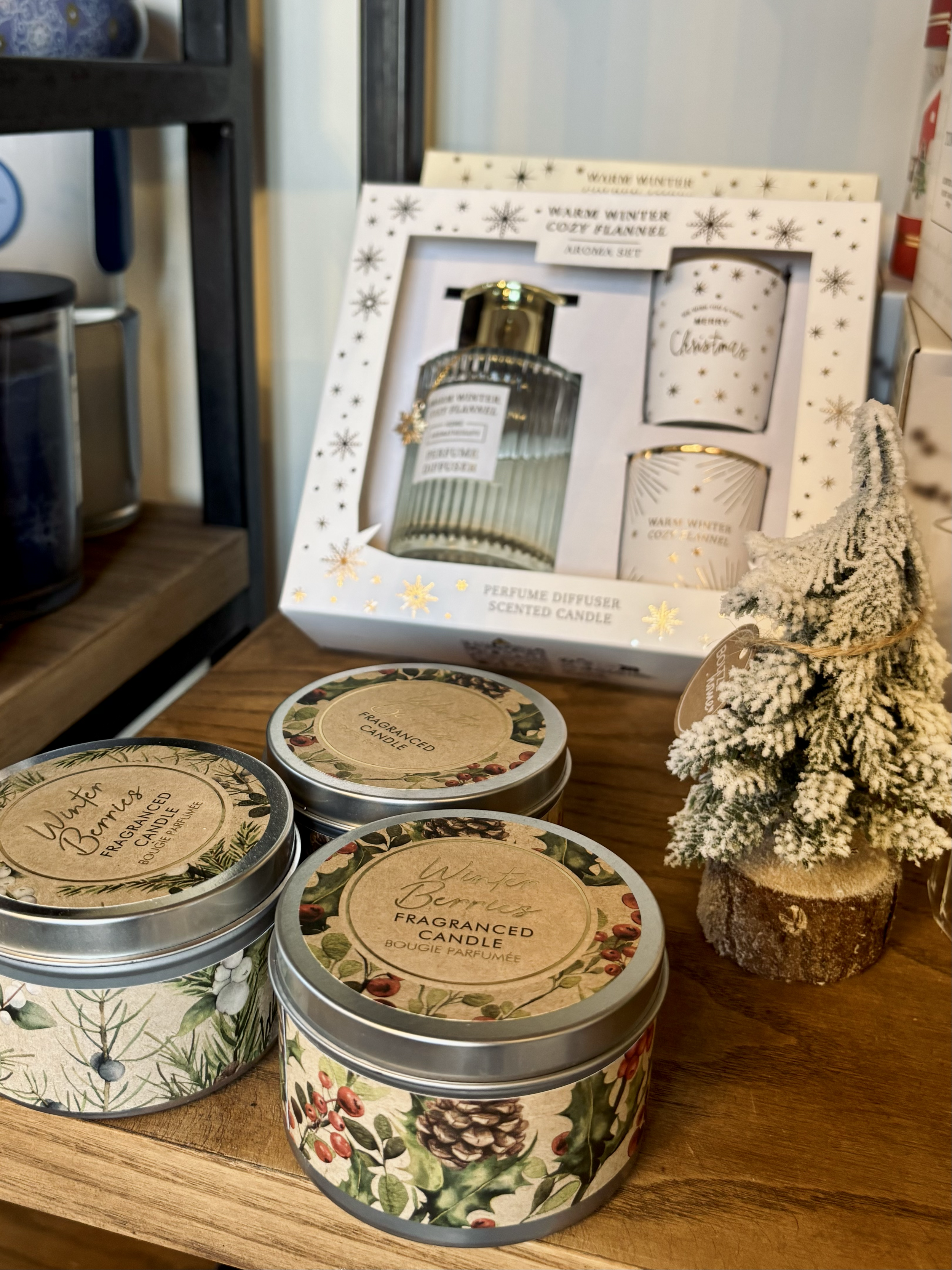 Set di prodotti natalizi decorativi con candele profumate, diffusore d'ambiente e un albero di Natale artificiale decorato, su un tavolo di legno.