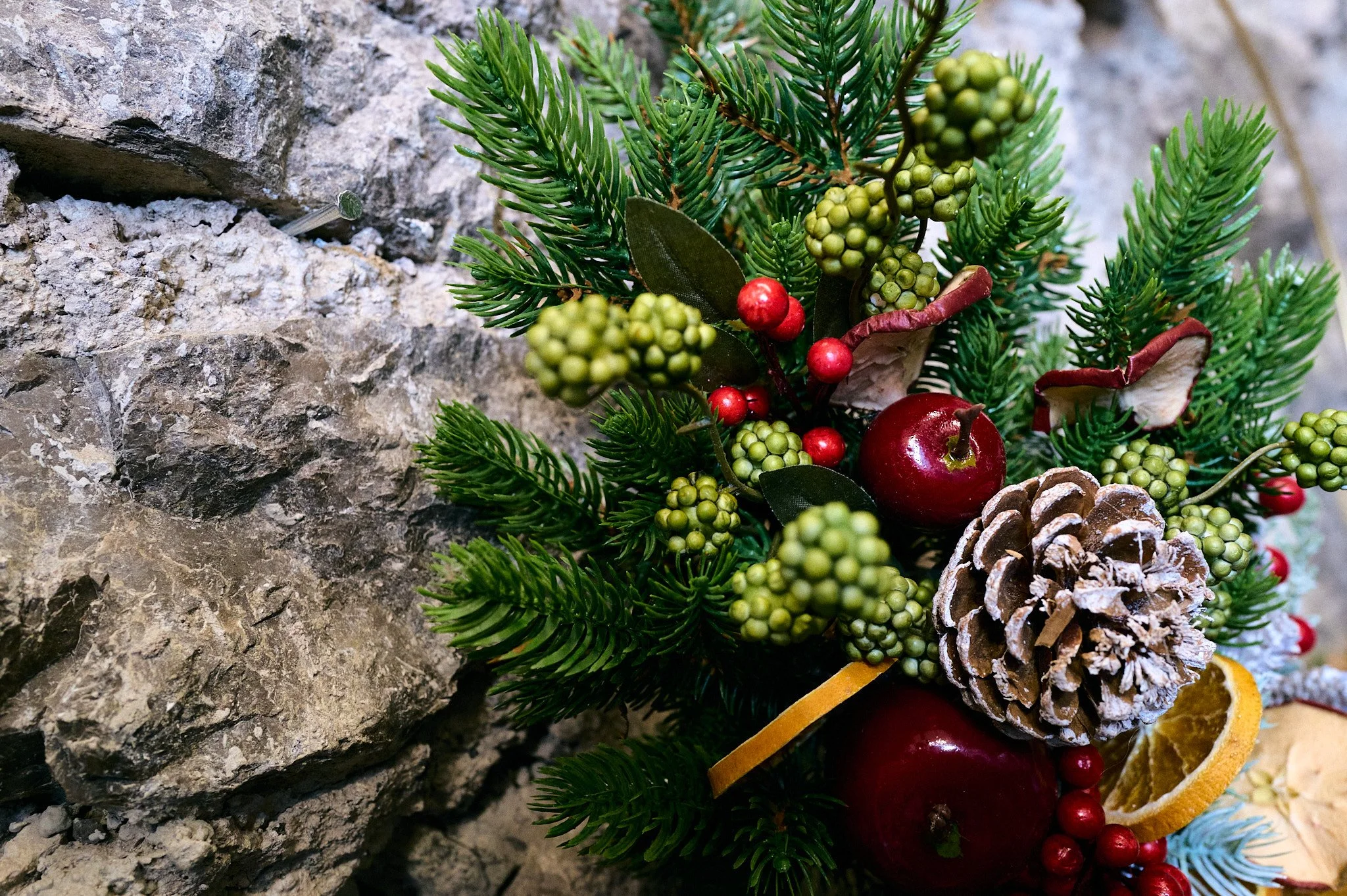 Ghirlanda natalizia con pigne, mele rosse, bacche rosse e foglie verdi ornamentali, decorata con frutta secca e agrumi, appoggiata su una superficie di pietra.