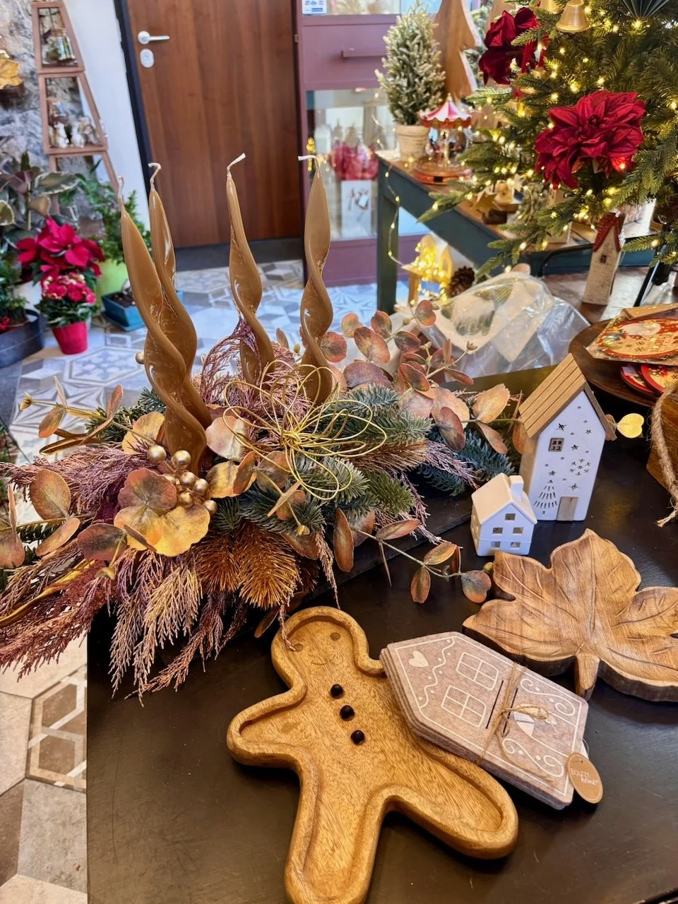  decorazioni natalizie su un tavolo, tra cui rami secchi, candele avvolte in filo di oro, statuine a forma di casa, un albero di Natale decorato, foglie d'acero in legno, un biscotto a forma di ombra, e altri ornamenti festivi.