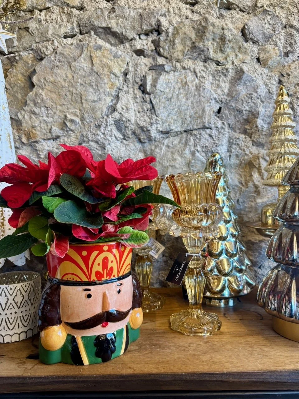 Vaso decorativo a forma di uomo con cappello rosso e barba nera, contenente un vaso di poinsettia rossa, circondato da decorazioni natalizie in vetro colorato e un albero di Natale in vetro riflettente.