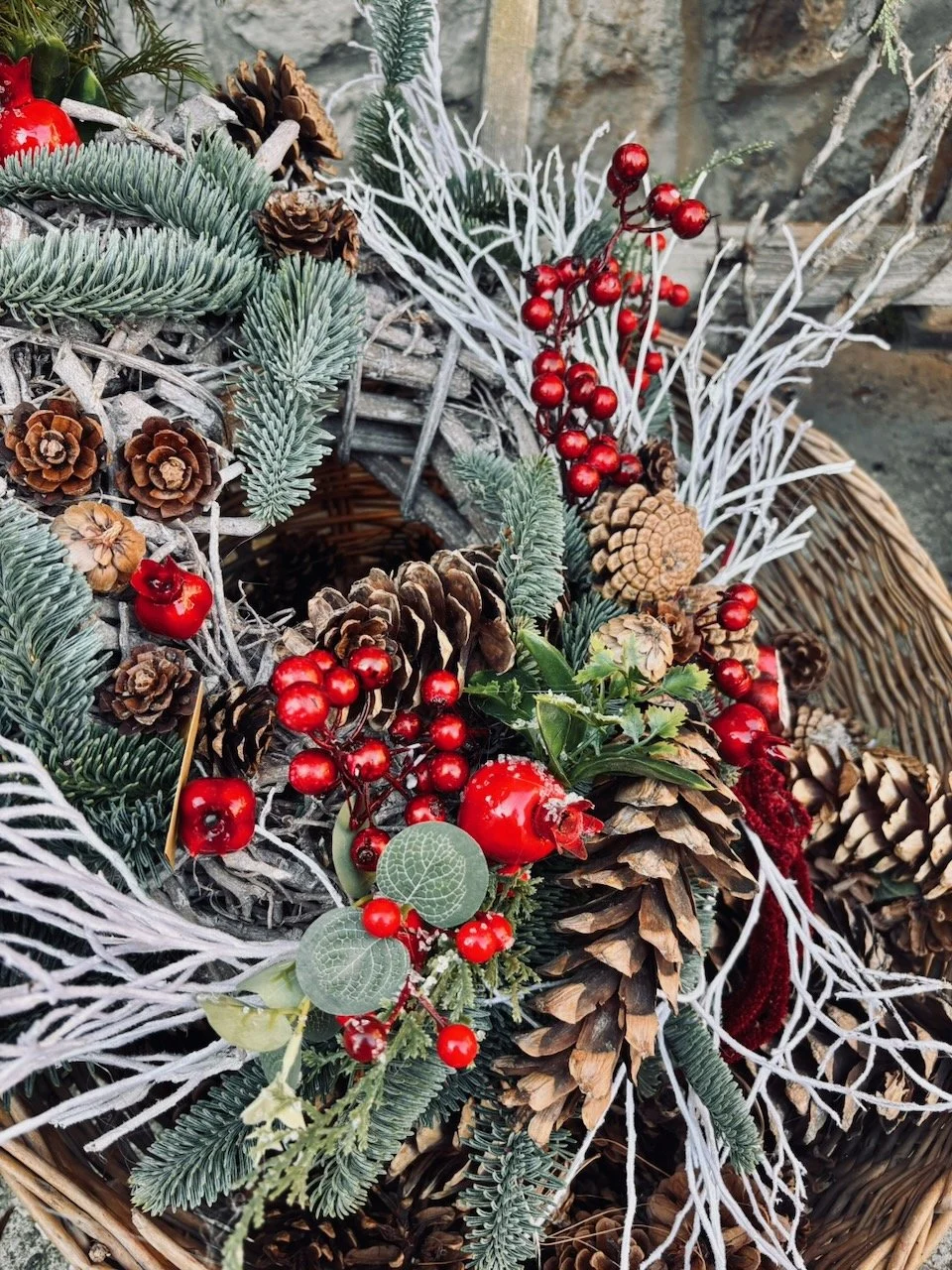 Corone natalizie decorata con pigne, aghi di pino, bacche rosse, e rami argentati, in un cesto di vimini.