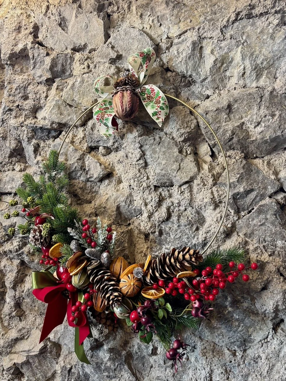 Decorazione natalizia a forma di cerchio con rami di pino, pigne, bacche rosse, agrumi essiccati, una goccia di fiocco rosso e verde, e un fiocco con motivo natalizio appeso a una parete di pietra.