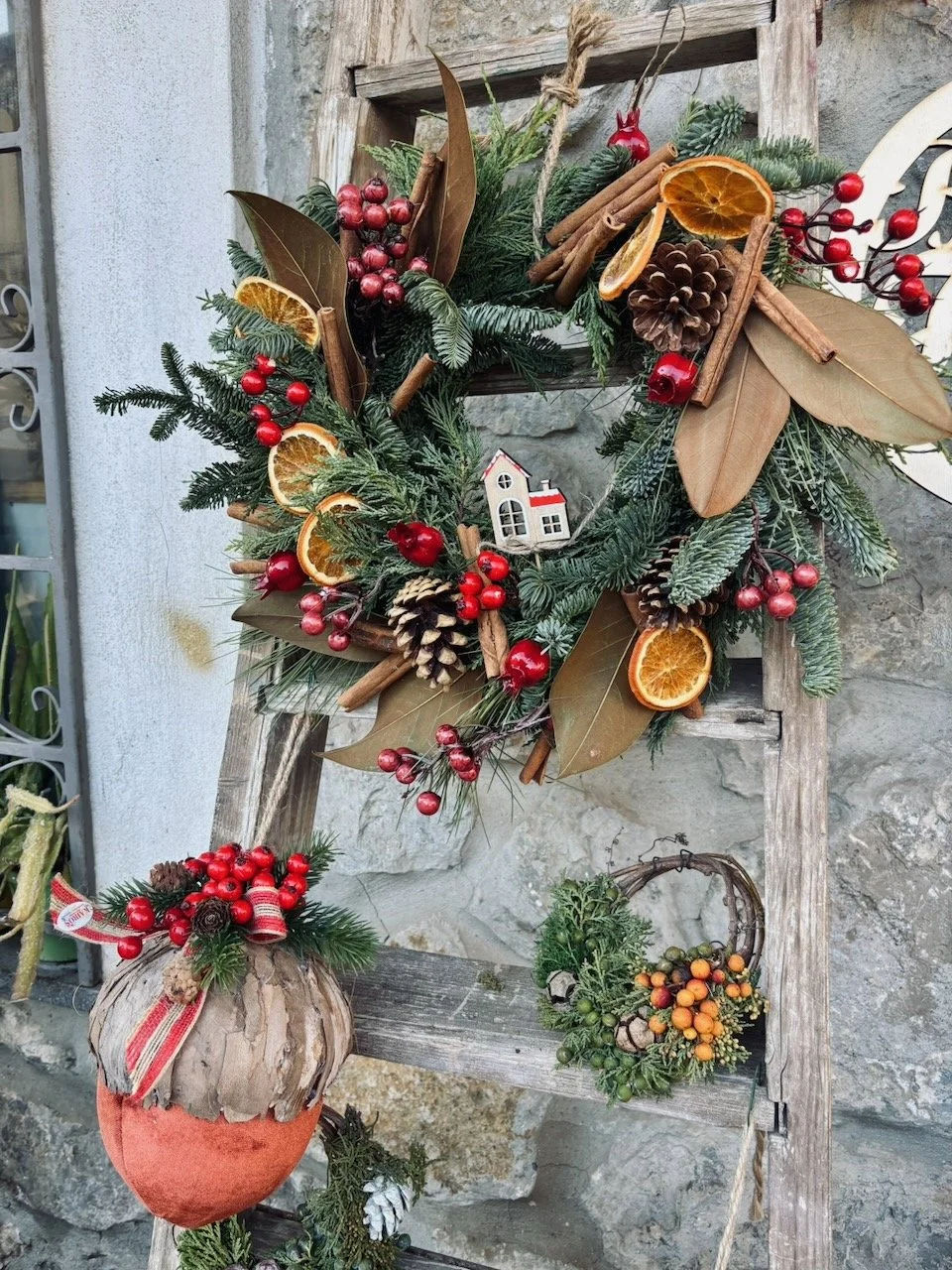 Corona dell'Avvento decorata con agrumi secchi, pigne, bacche rosse, cannella e foglie di edera, appesa a una struttura di legno, con vasi di piante e decorazioni natalizie.