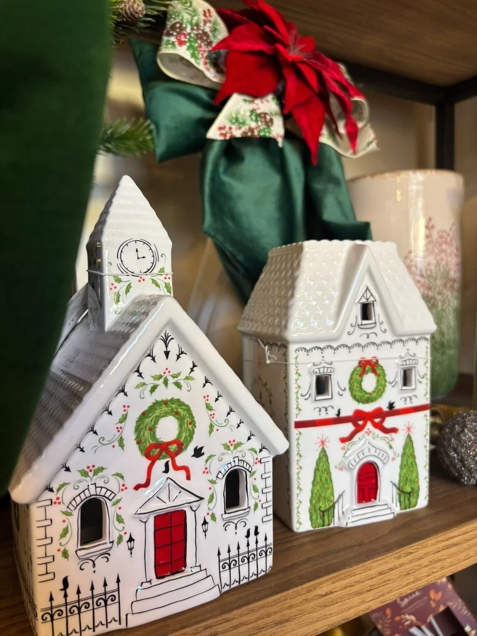 Decorazioni natalizie con case di ceramica bianche decorate con motivi natalizi, ghirlande verdi e nastri rossi, su una mensola di legno.