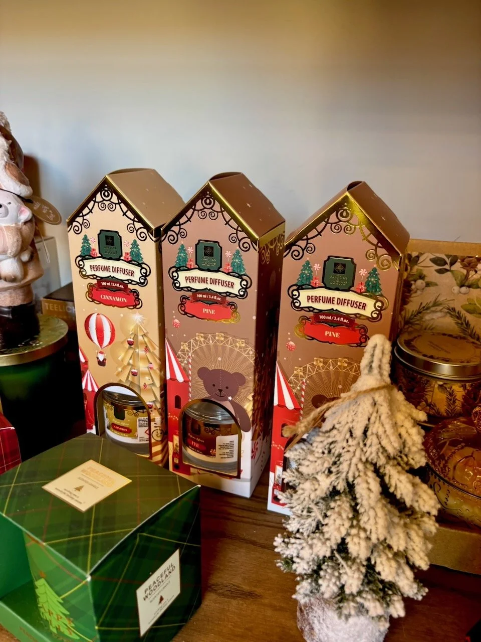 Tre confezioni di profumo diffusore in stile natalizio, decorati con un albero di Natale e un orsetto, con una piccola pianta finta innevata accanto.