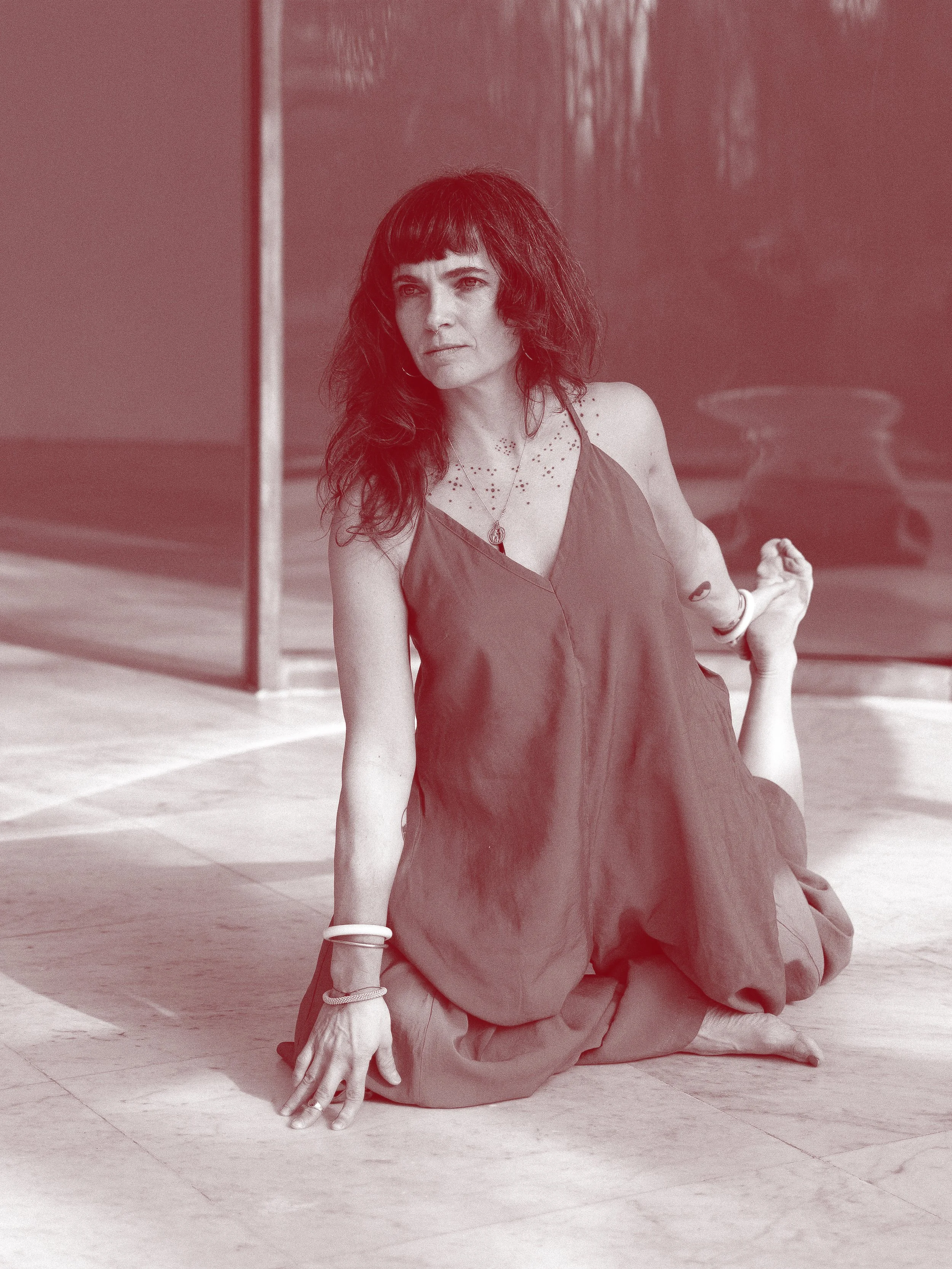Eine Frau sitzt auf dem Boden in einer Yoga-Position, die linke Hand berührt den Boden, die rechte Hand ist auf dem Knie, blickt nach vorne, trägt ein dunkles Kleid, einige Armreifen, ein Halskettchen, im Hintergrund eine Pflanze und eine Steinskulptur.