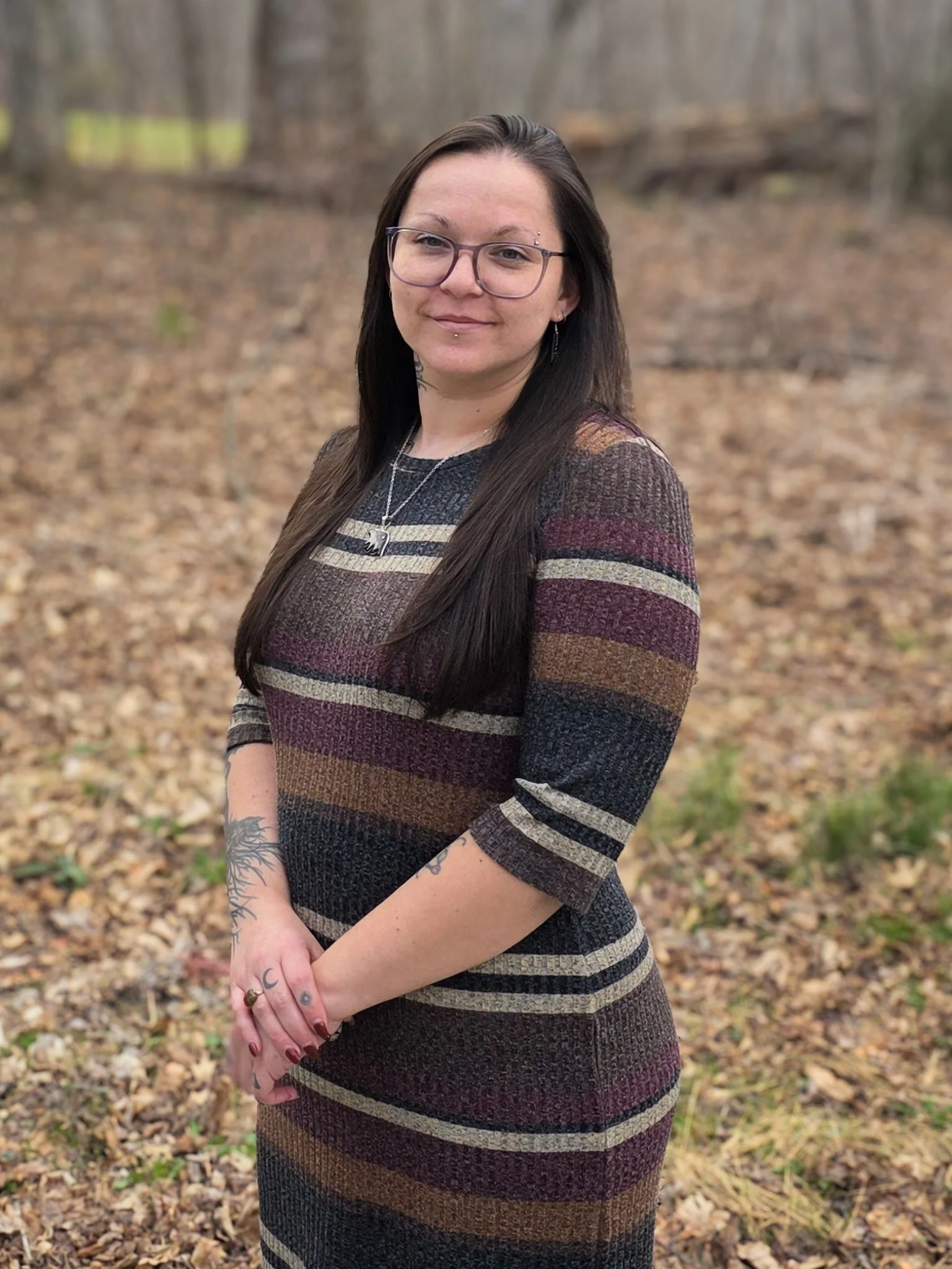 A woman with long dark hair and glasses standing outdoors in a wooded area with fallen leaves on the ground.
