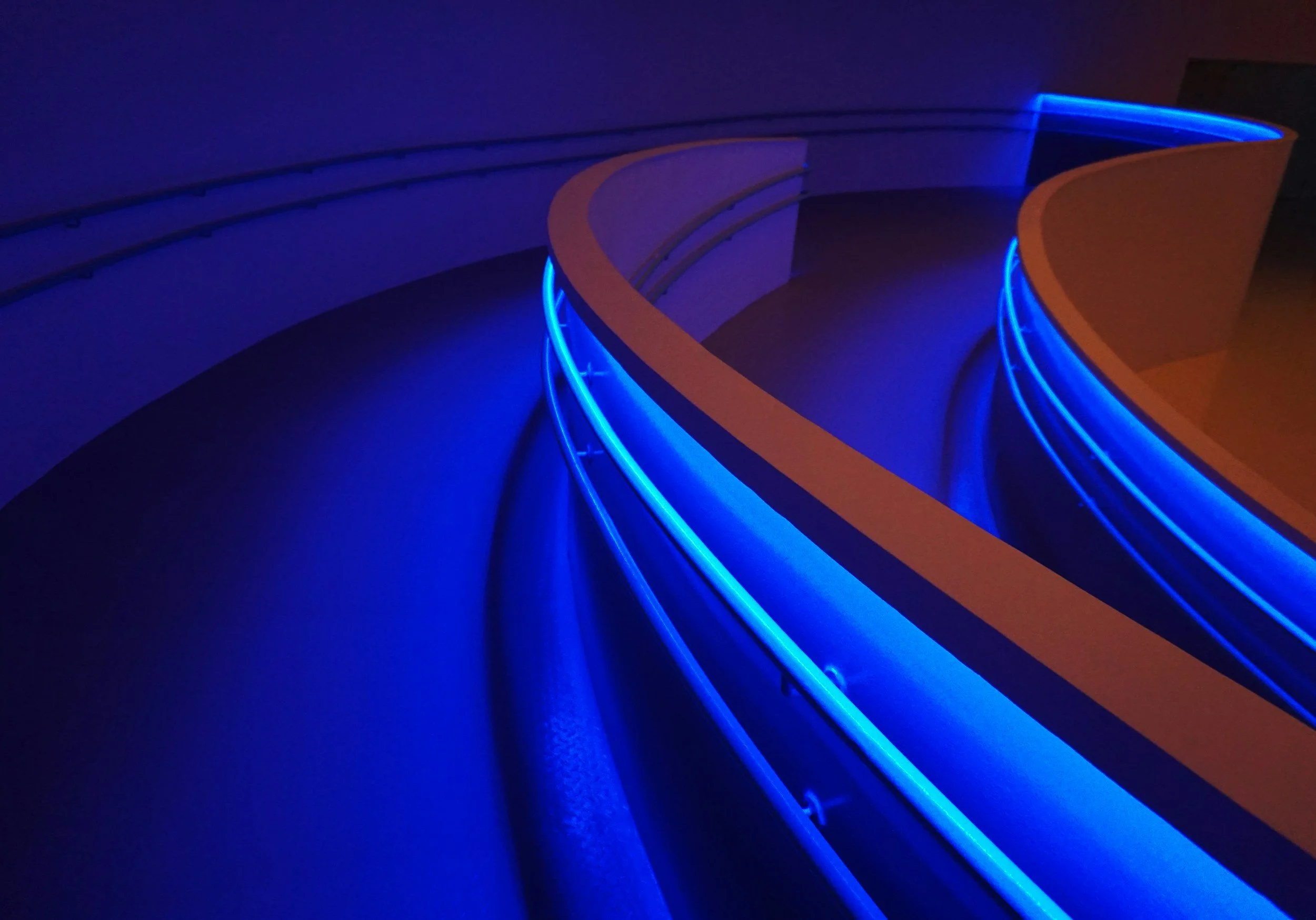 Futuristic curved corridor with blue neon lights along the railings and orange walls, dimly lit.