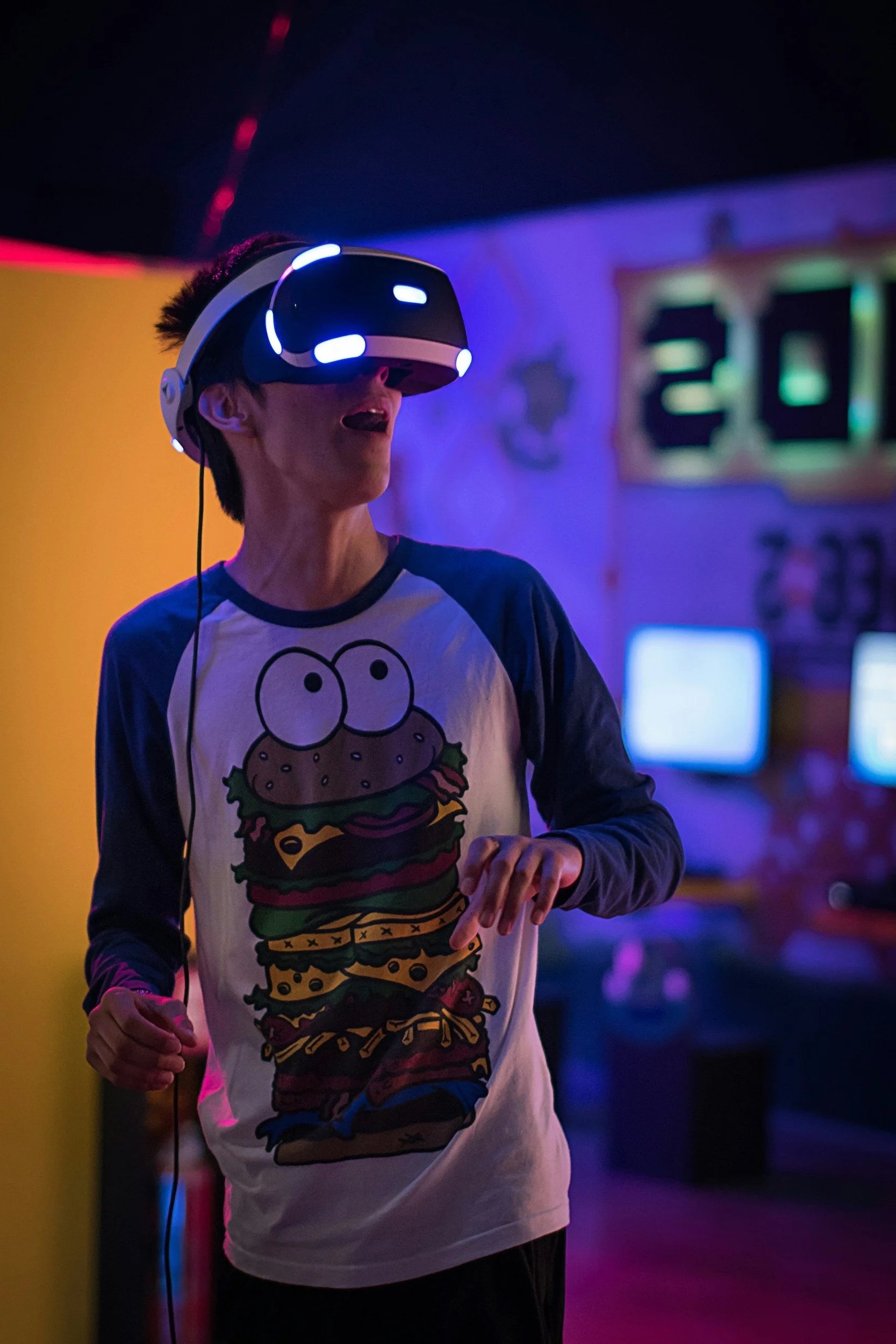 A young man wearing a virtual reality headset in a game arcade or entertainment center, with colorful neon lighting in the background.