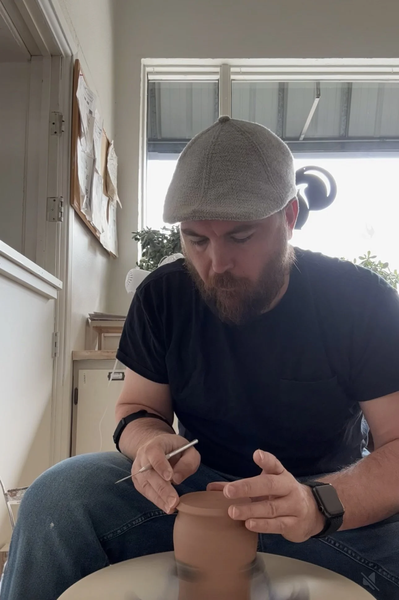 A man with a beard, wearing a flat cap and black shirt, is seated at a pottery wheel, shaping a clay pot with a sculpting tool. He is indoors with plants and bulletin boards in the background.