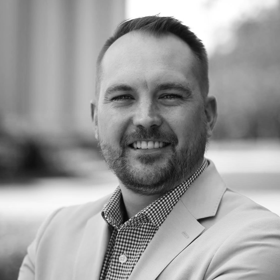 Black and white portrait of a smiling man with short hair, a beard, wearing a blazer over a checked shirt, outdoors with blurred background.