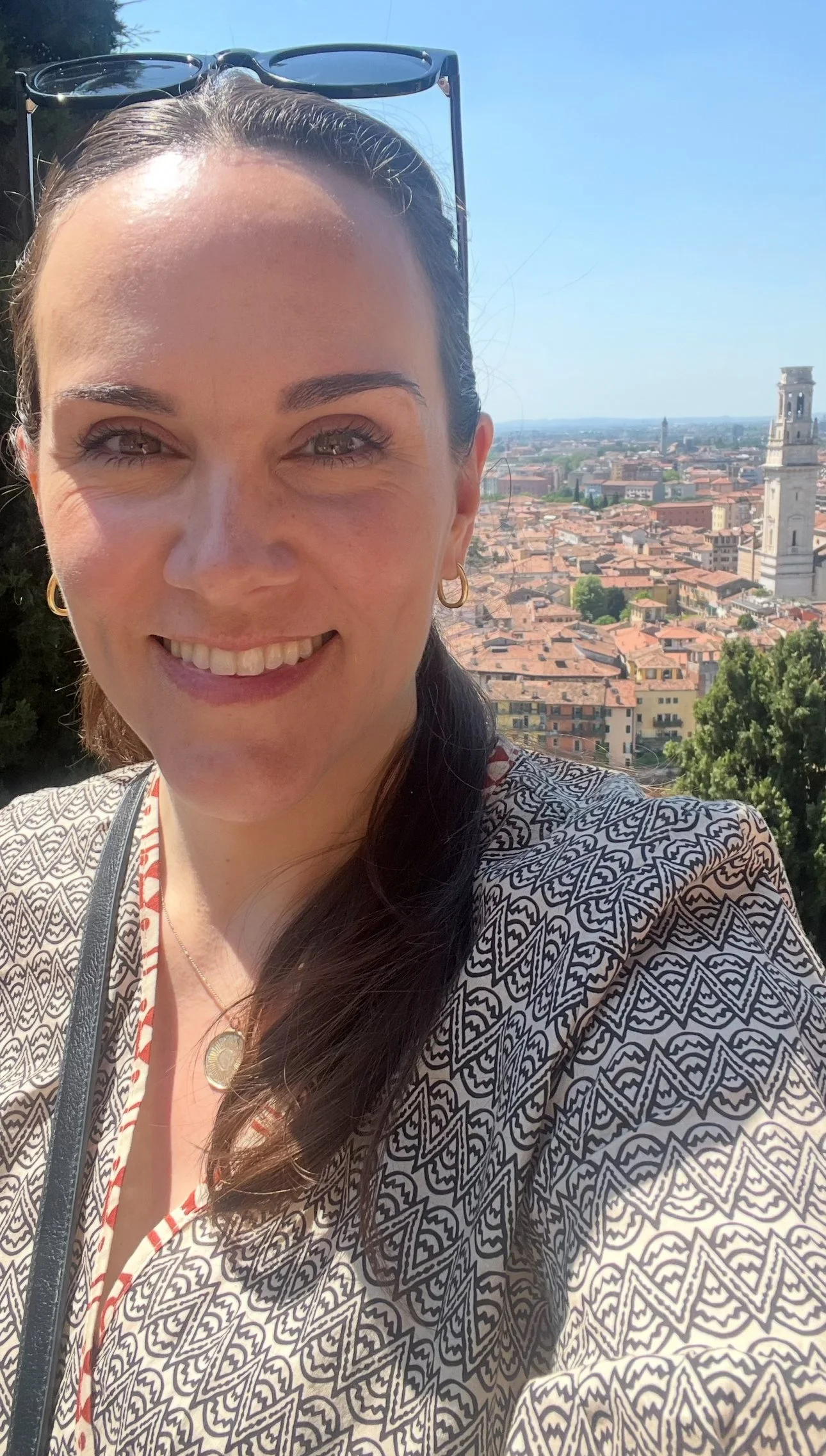 A woman smiling outdoors with a cityscape in the background, including red rooftops and a tall clock tower, under a clear blue sky.