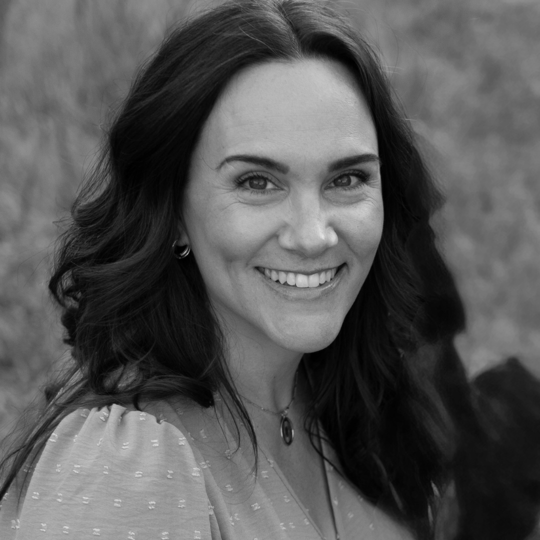 Black and white portrait of a smiling woman with wavy dark hair, wearing earrings and a necklace, outdoors.
