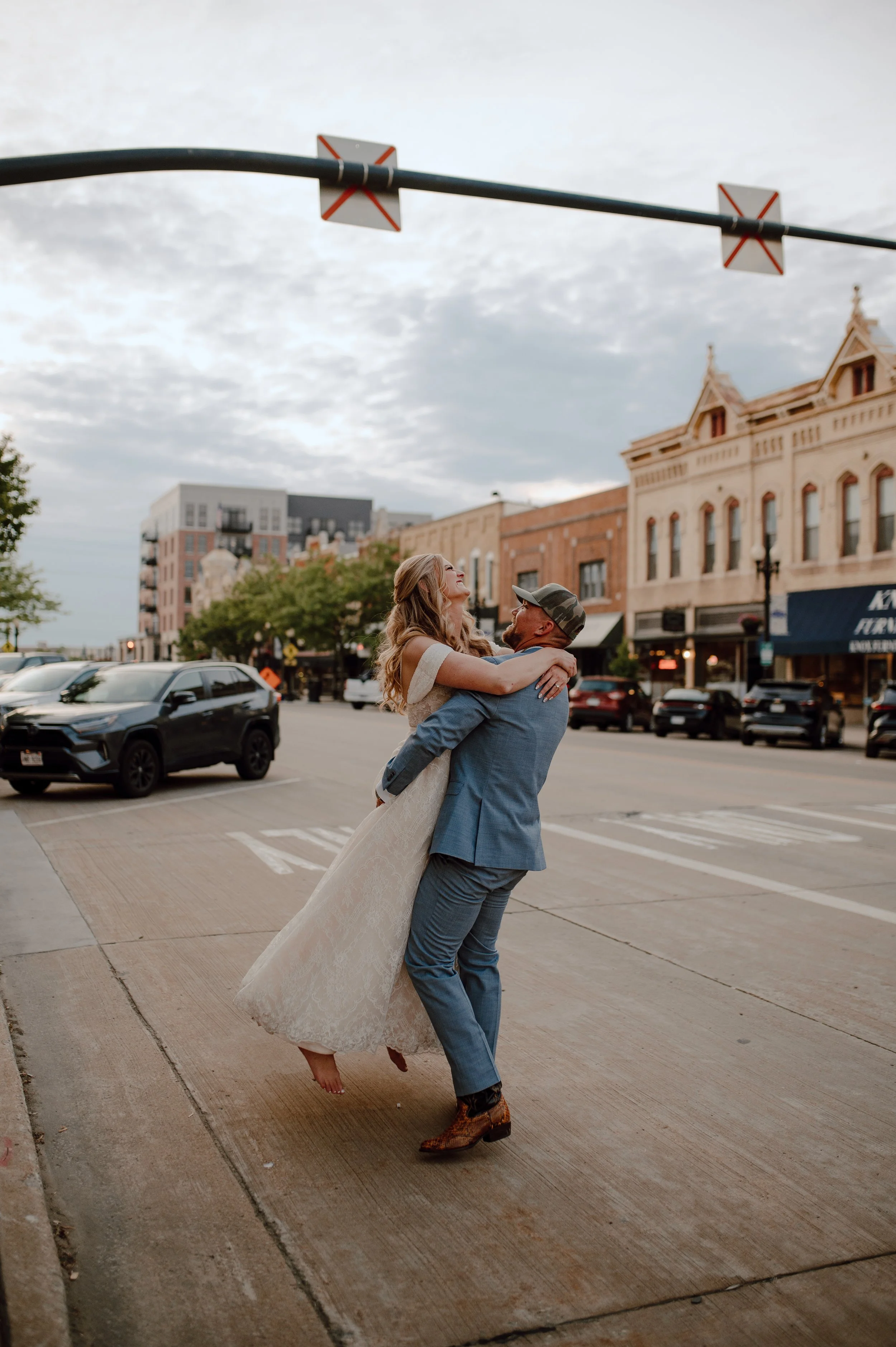 A man in a blue suit and a woman in a wedding dress dancing on a city sidewalk, with cars and historic buildings in the background on a cloudy day.