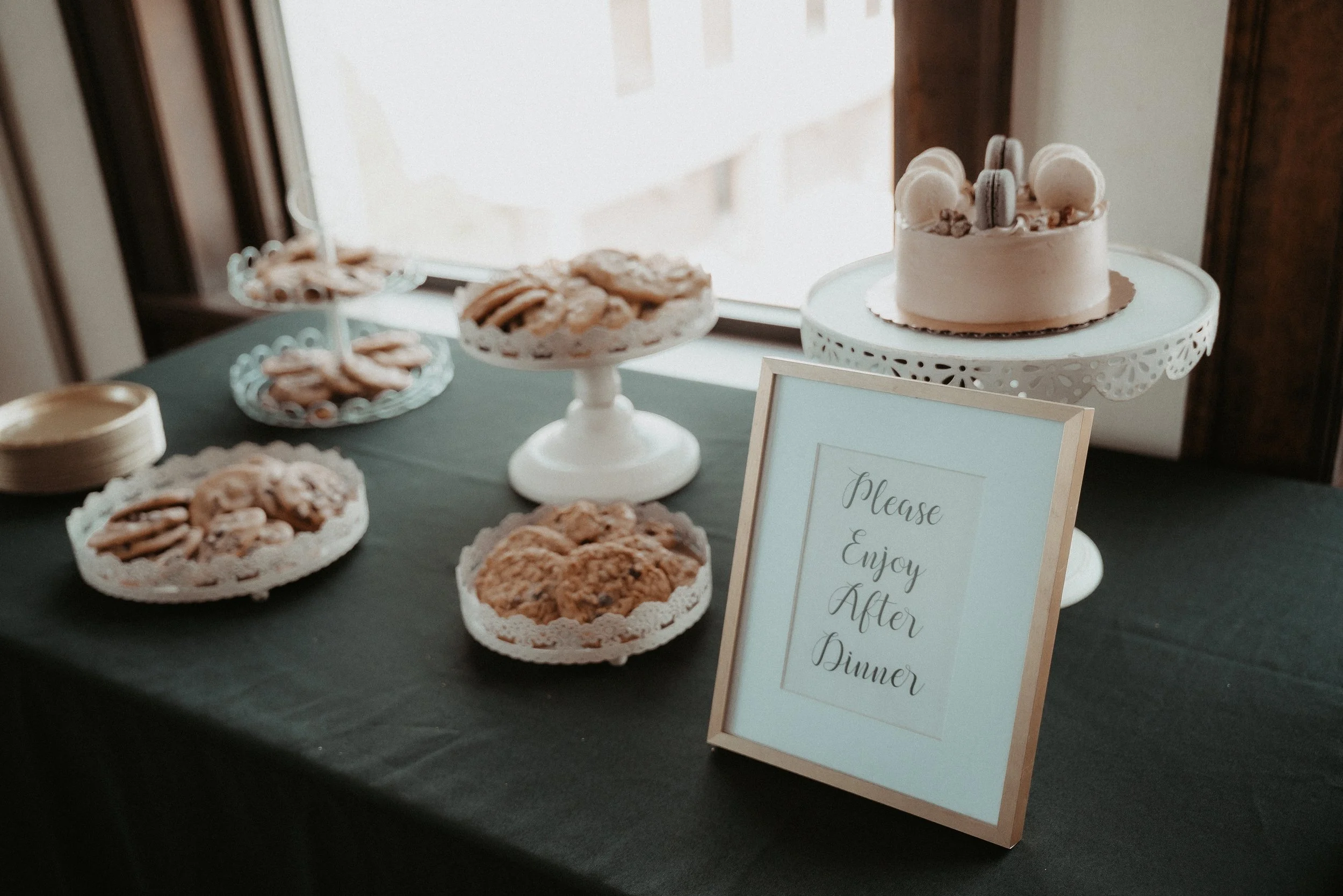 A dessert table with a white cake decorated with macarons and flowers, several plates of cookies, and a sign that says 'Please Enjoy After Dinner'.