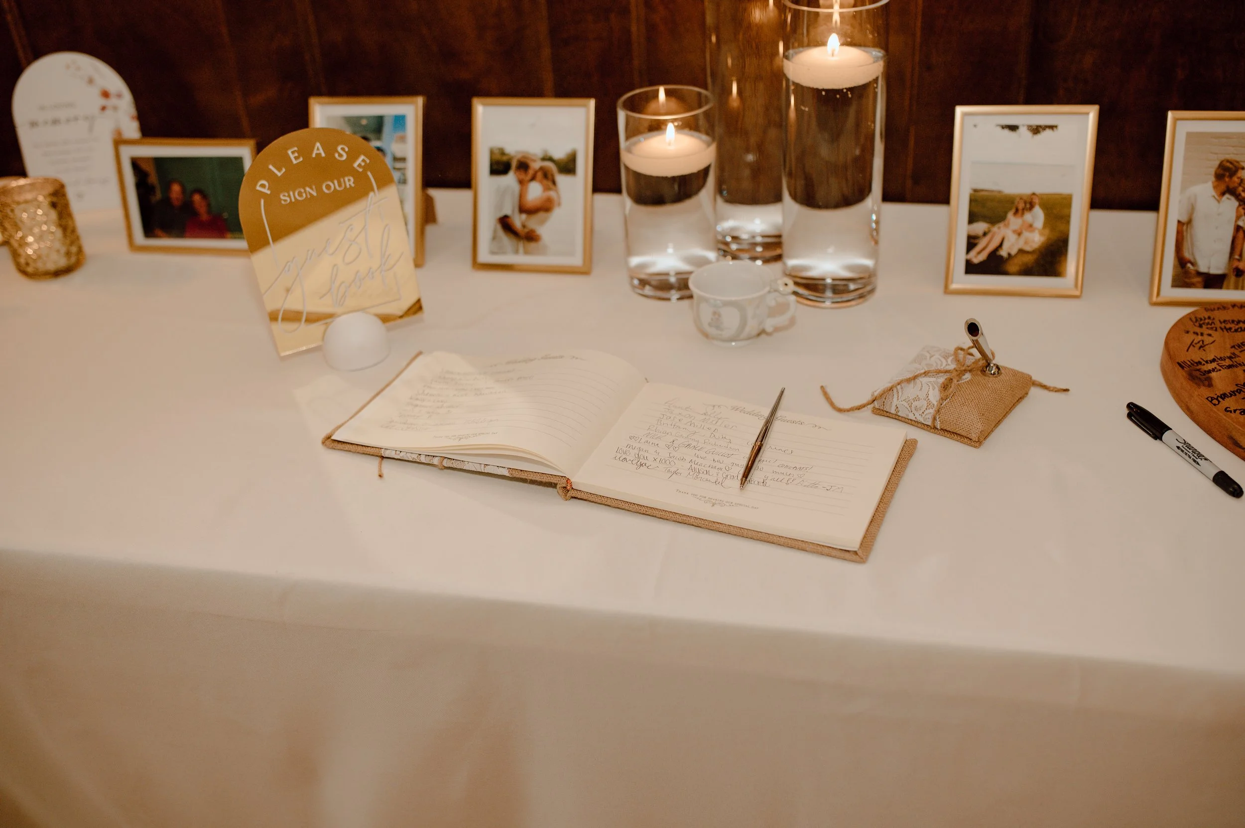 A guestbook on a wedding reception table with a pen, informational signs, framed photographs, lit candles, a small cup, and decorative items.