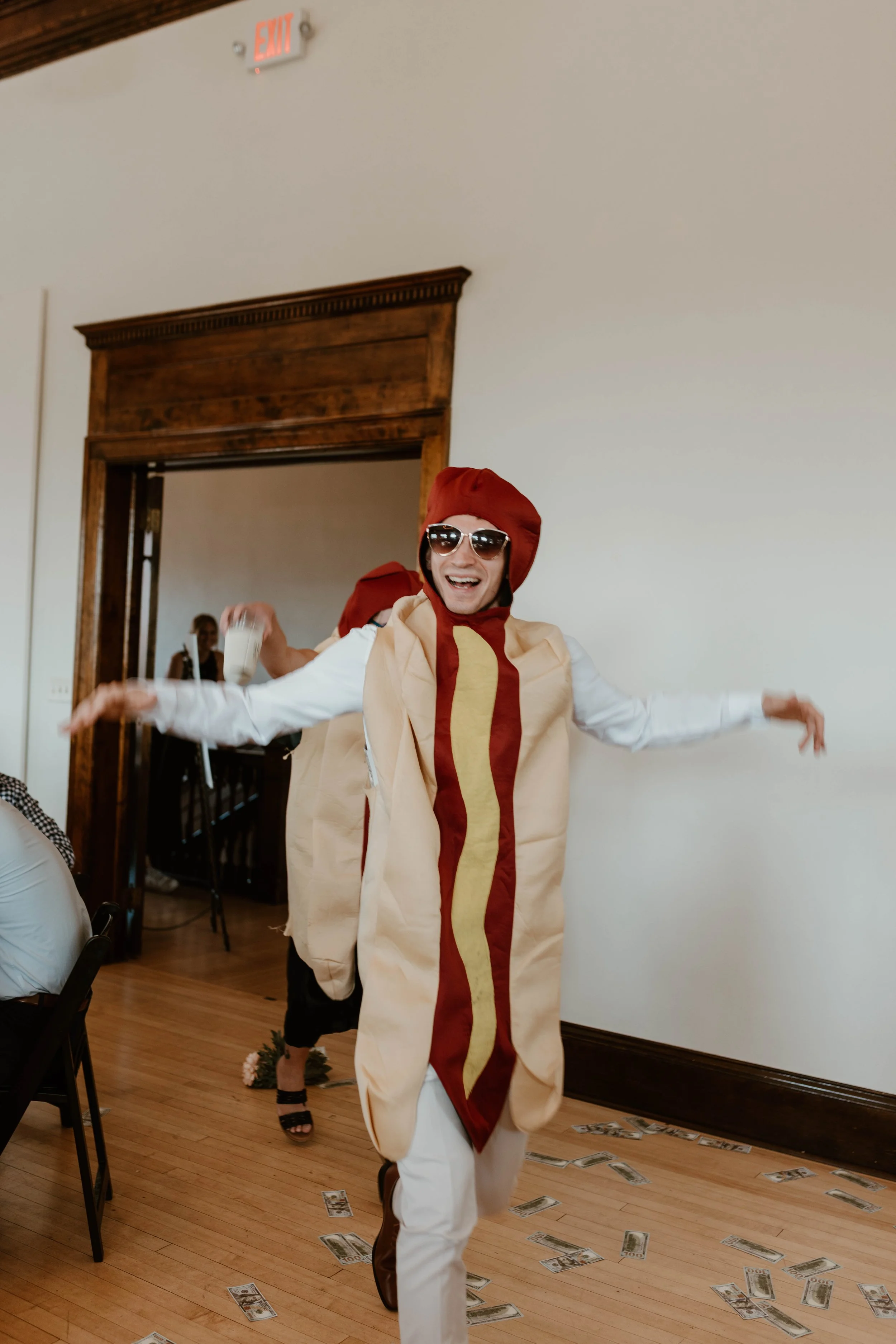 Person dressed as a hot dog with a bun, mustard, and ketchup, wearing sunglasses, dancing joyfully indoors with people and dollar bills on the floor.