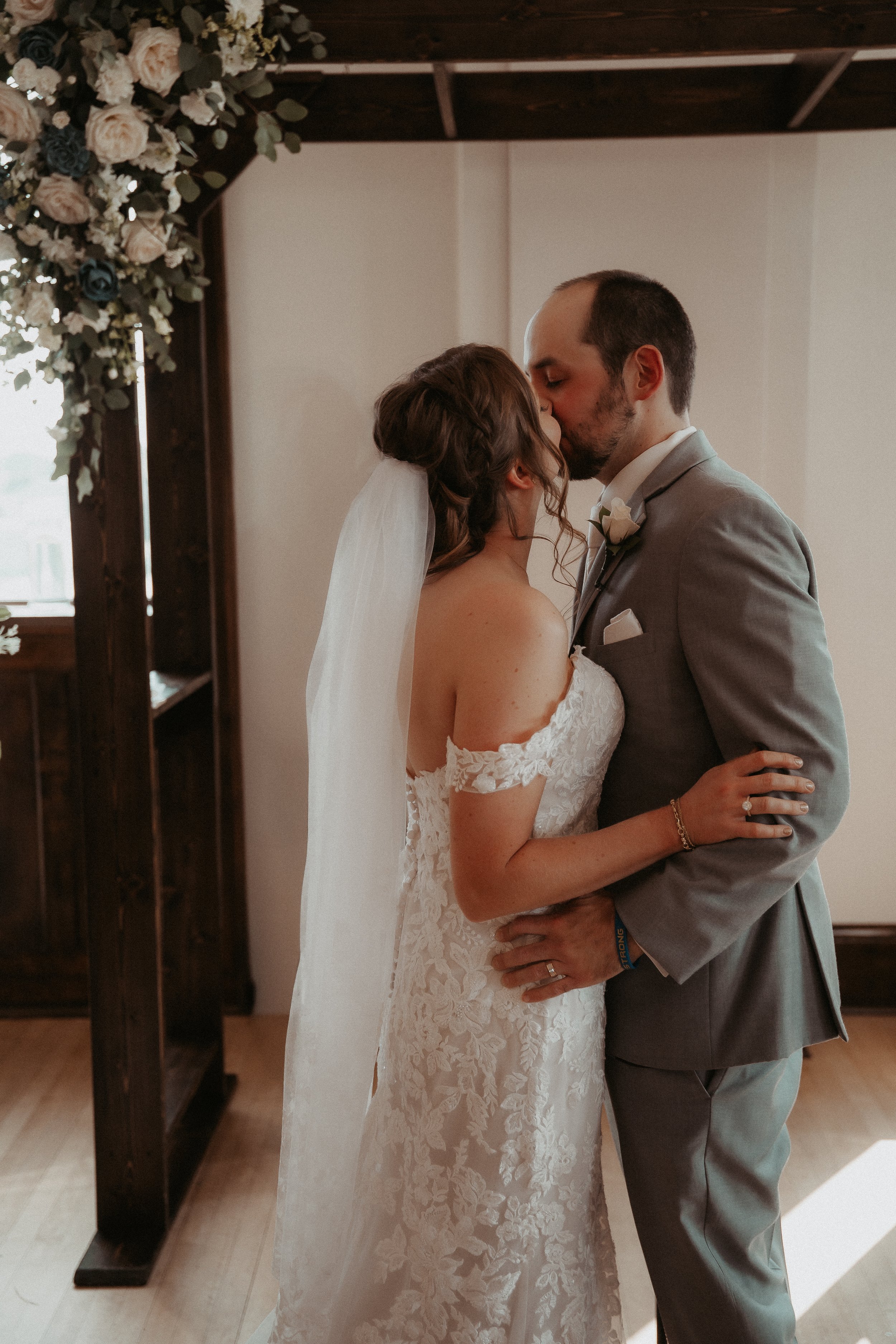 A bride and groom kissing indoors at their wedding ceremony, with floral decorations nearby.