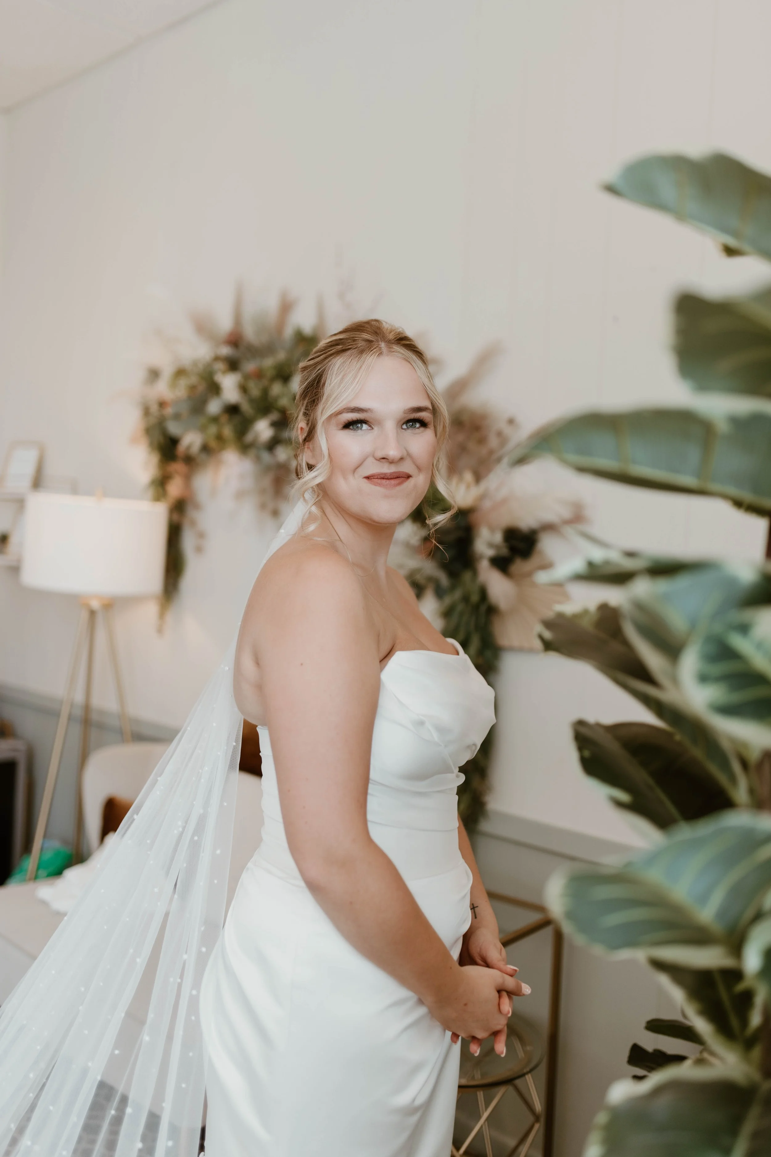 A smiling bride stands in a softly lit room with a floral backdrop and modern decor, wearing a strapless white wedding gown with a veil.