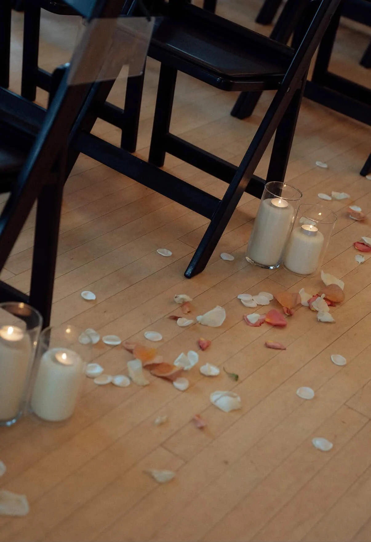 Decorative setting on wooden floor with scattered rose petals, white candles in glass holders, and empty black chairs