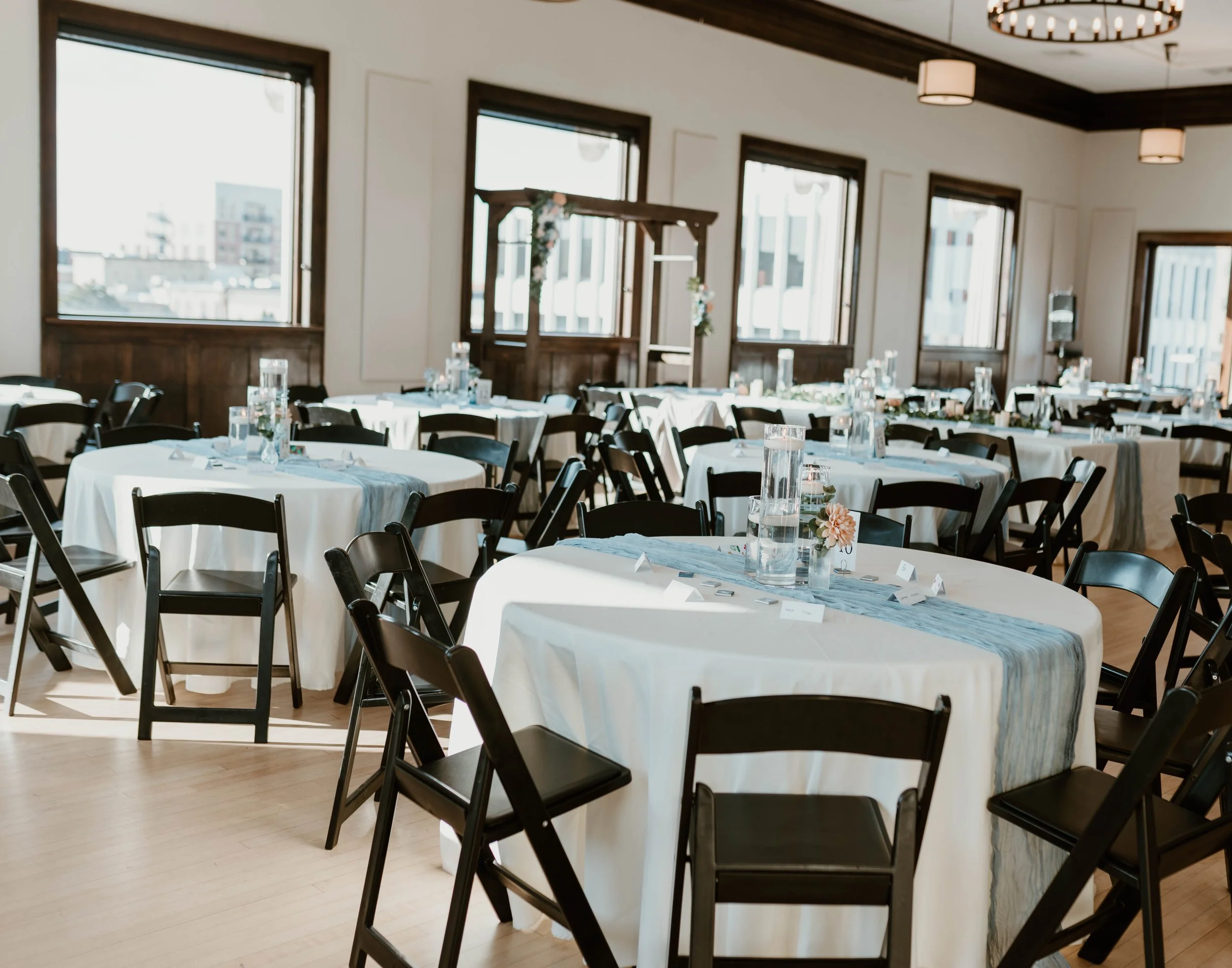 A decorated event space with round tables covered in white tablecloths, adorned with centerpieces and water pitchers, set for a celebration or gathering.
