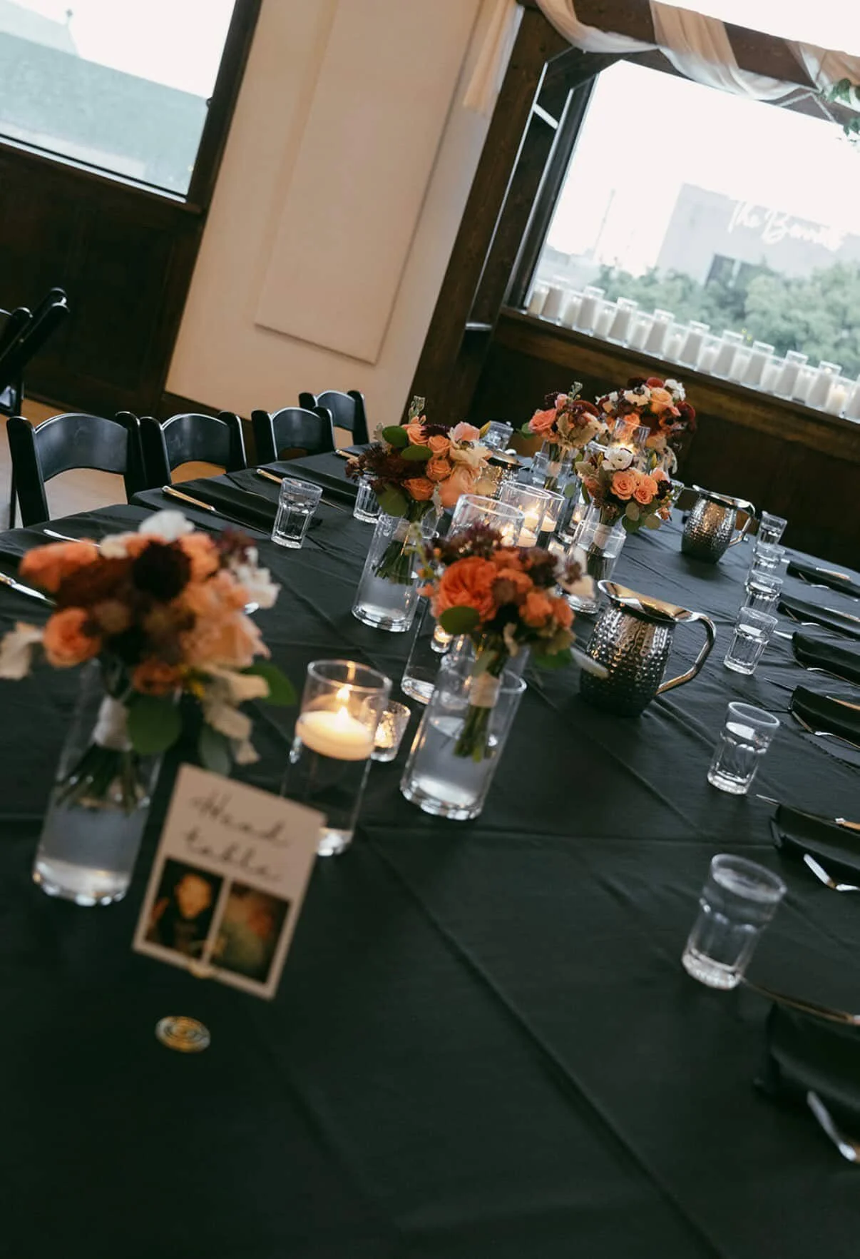 Elegant indoor banquet table set with black tablecloth, floral centerpieces, candles, and glassware, with windows showing an outdoor view.