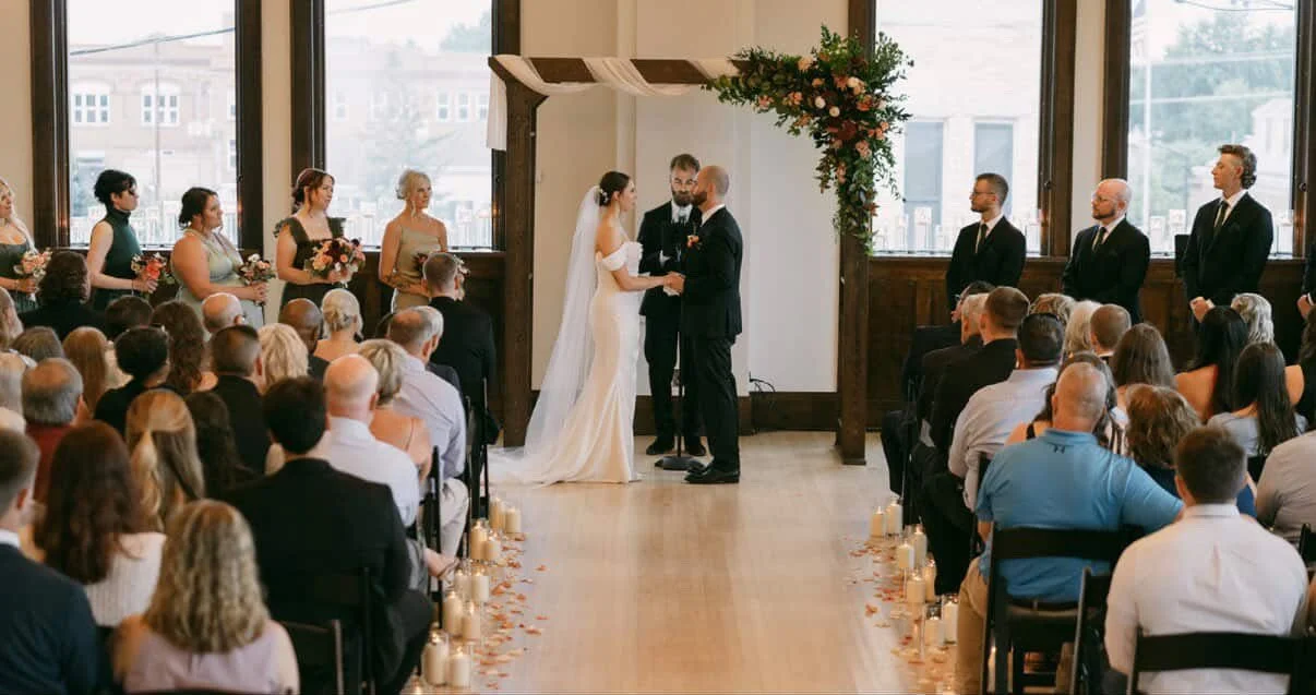 A wedding ceremony with a bride and groom holding hands, exchanging vows in front of officiant, in a bright room with large windows. Bridesmaids and groomsmen stand on either side, guests seated in rows, candles and flower petals decorate the aisle.