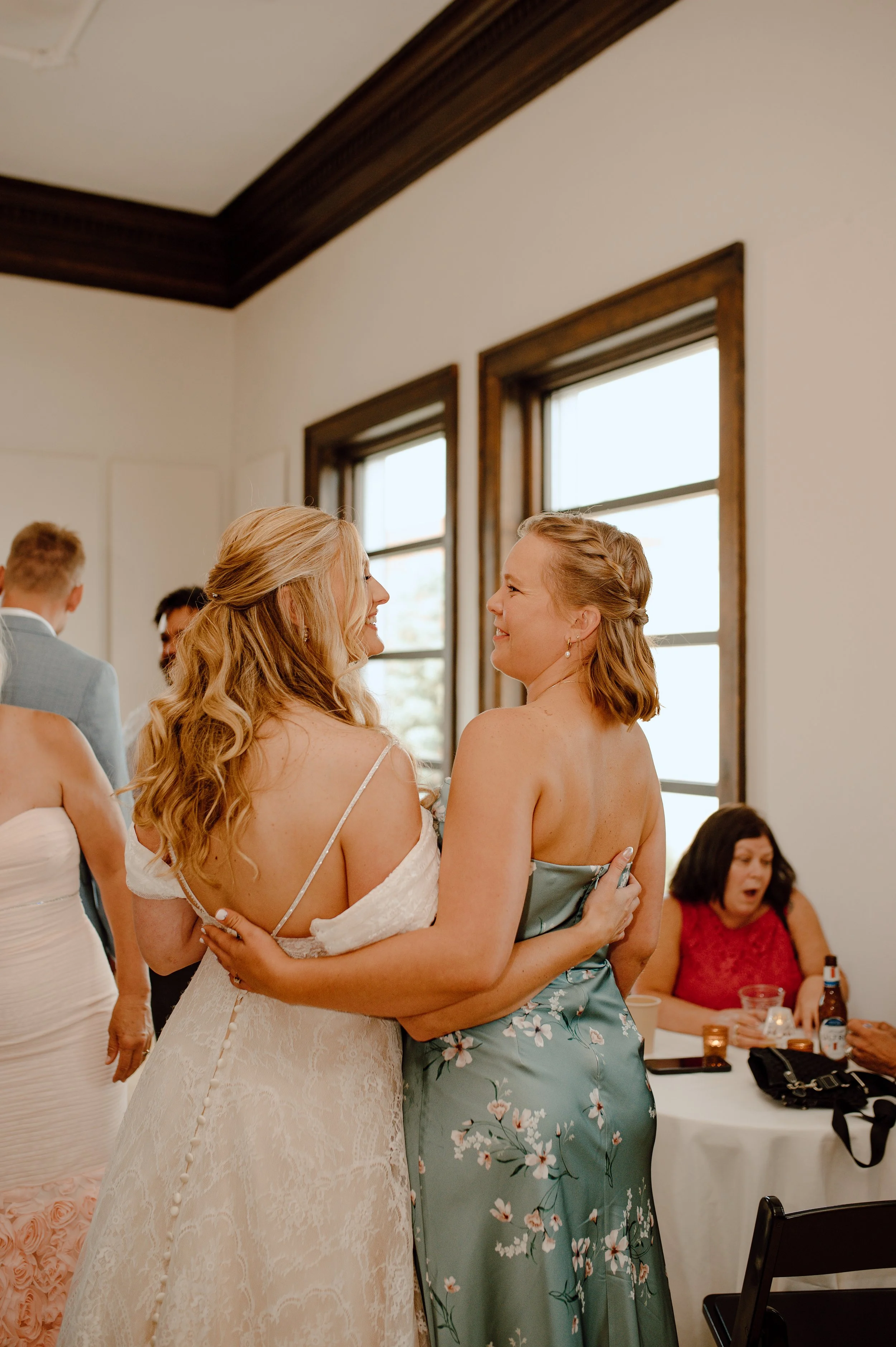 Two women embracing and smiling at each other during a wedding celebration, with other guests in the background.