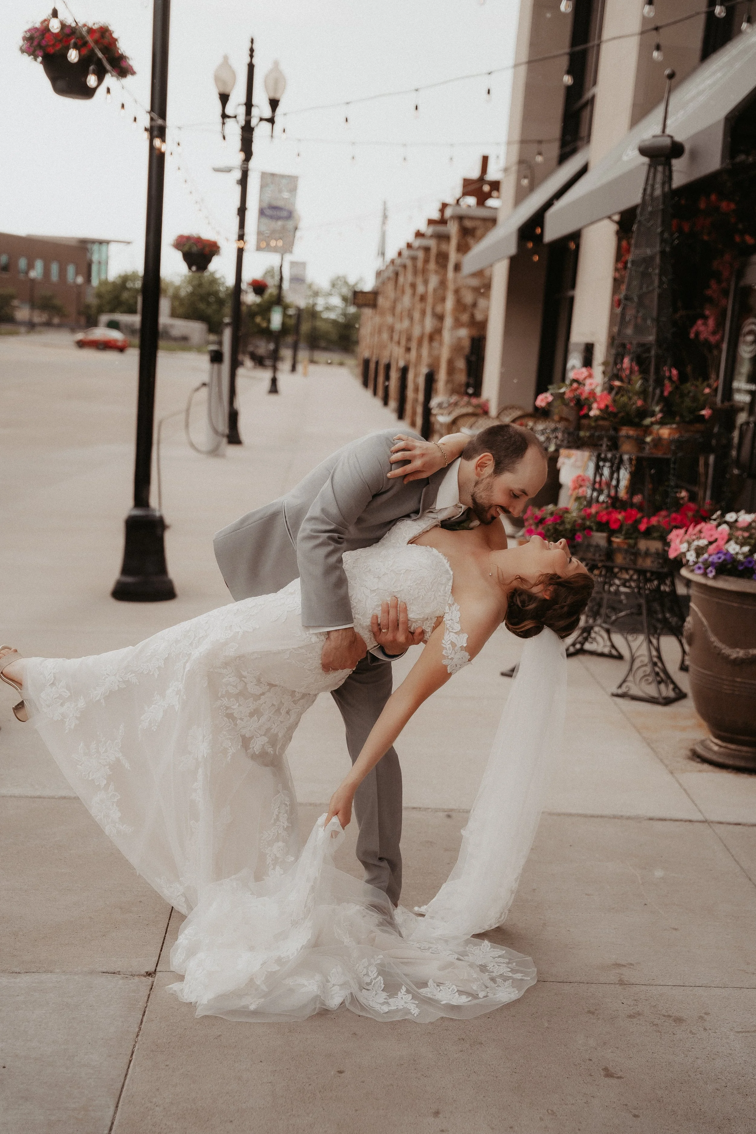 A groom dipping and kissing a bride in a romantic pose on a sidewalk, with streetlights and storefronts in the background.