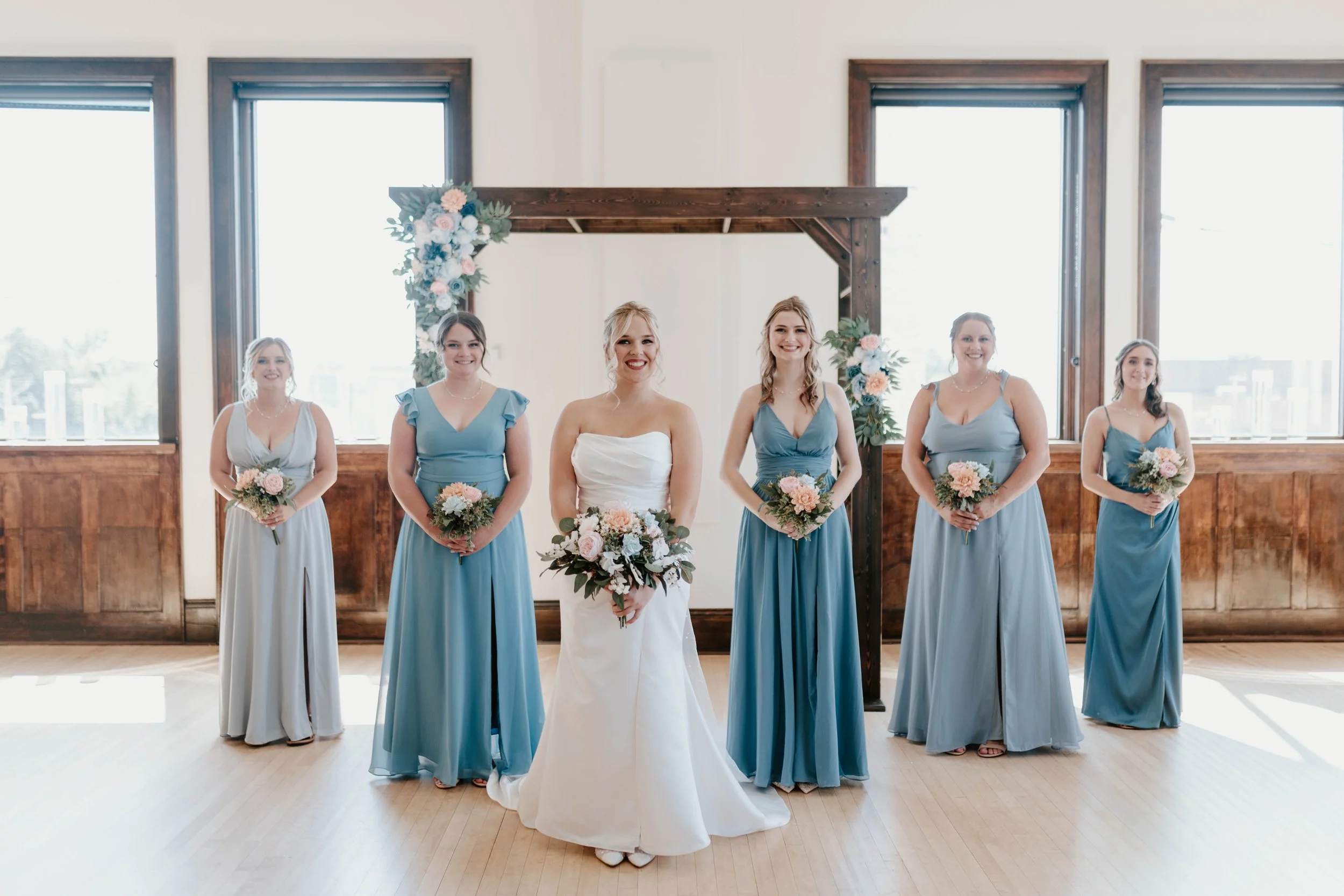 Bride in white wedding gown with five bridesmaids in light blue dresses standing in front of a wedding arch decorated with flowers, indoors with wooden paneled walls and large windows.