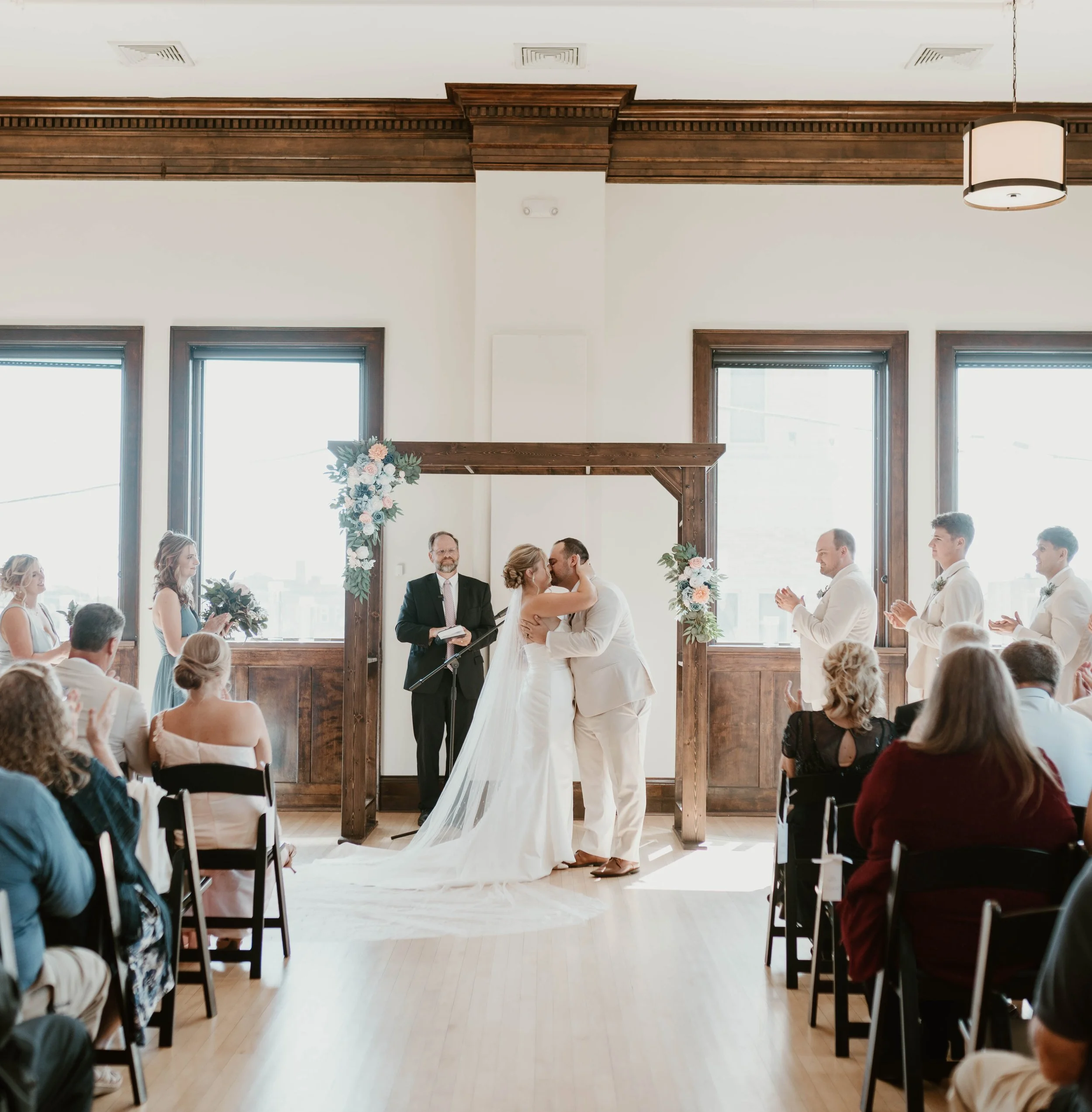 Bride and groom kissing during wedding ceremony under a wooden arch decorated with flowers, with officiant and guests applauding in a bright, spacious room with large windows.