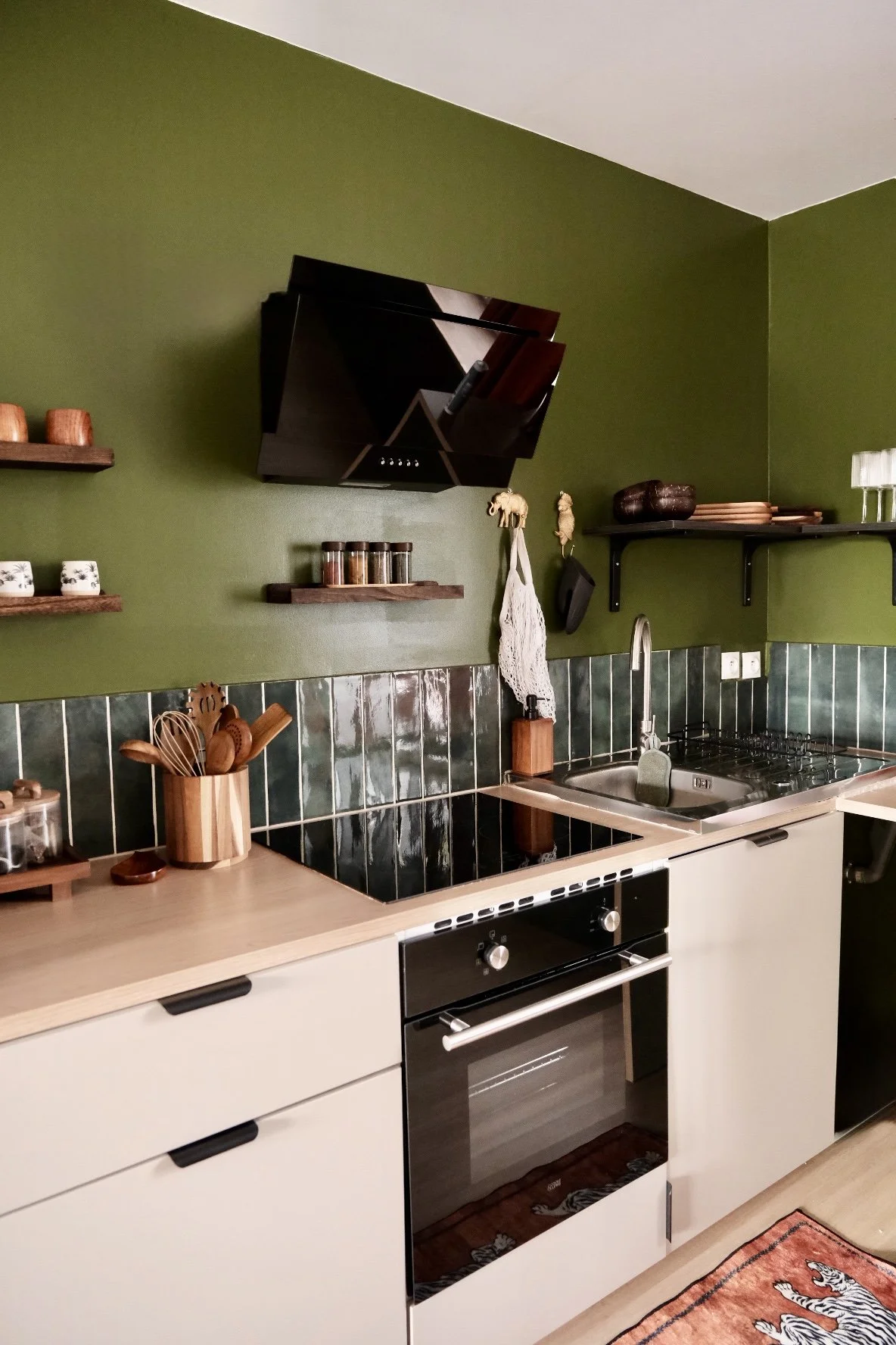 Cuisine moderne avec mur vert, étagères en bois, plan de travail en bois clair, four intégré, évier en acier inoxydable, accessoires en bois et décoration verte.