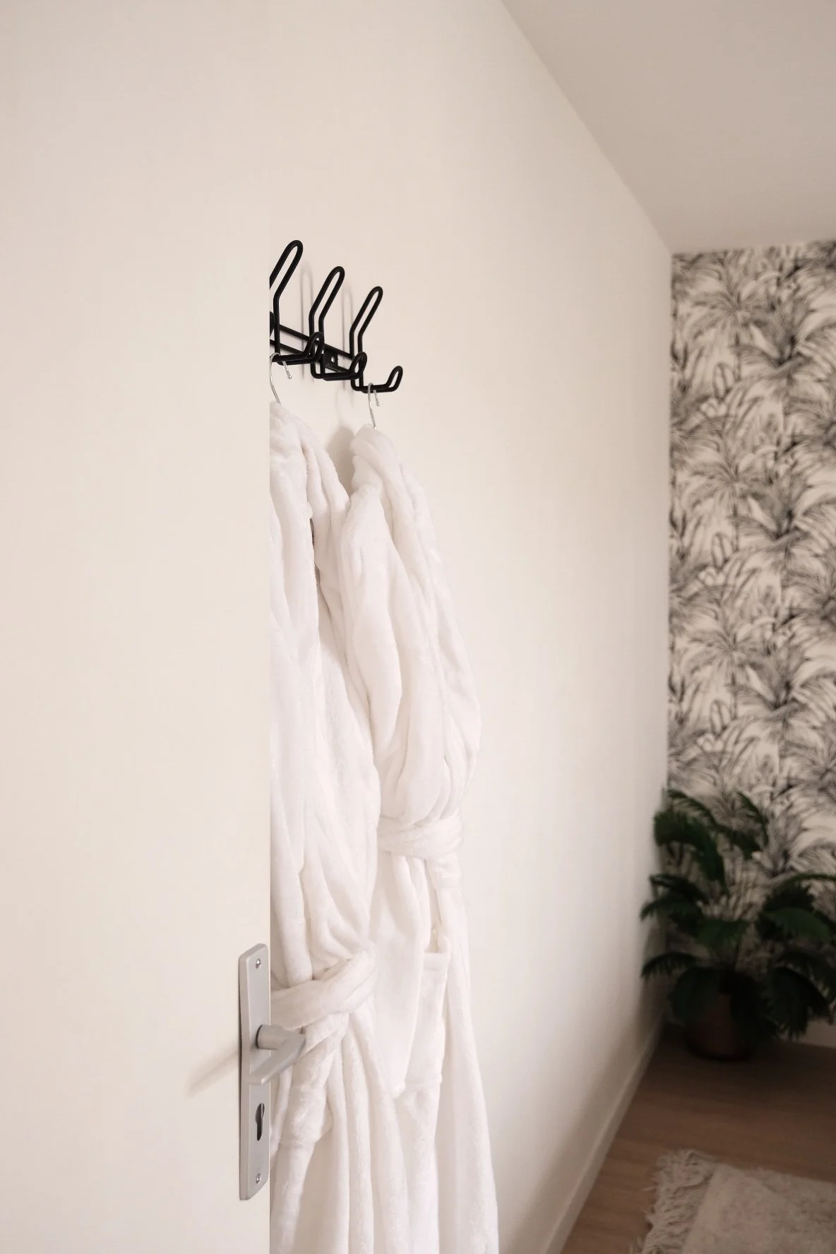 Peignoir blanc suspendu sur un cintre noir fixé au mur dans une pièce élégante avec papier peint floral en arrière-plan et une plante verte dans un pot en argile.