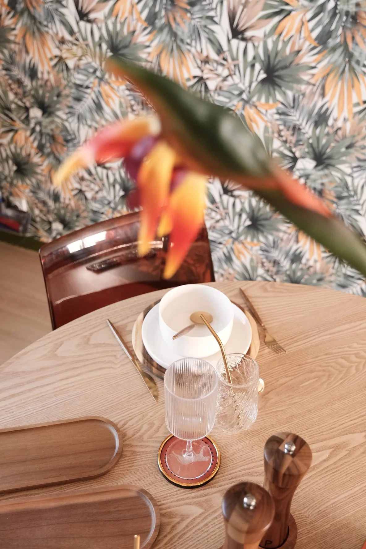 Une table en bois avec un verre à vin transparent, une assiette blanche, une cuillère en or, une fourchette, un chiffon de table en tissu beige, et une jardinière avec une plante à fleurs orange et jaune suspendue au-dessus de la table. En arrière-plan, un mur à motifs végétaux en vert, jaune et blanc.