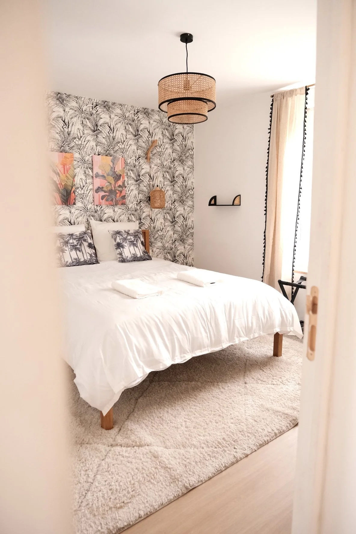 Chambre avec mur à motif tropical, lit avec draps blancs, oreillers à motif palmier, lampadaire en rotin, étagère noire, rideaux beige, tapis beige, décoration naturelle.