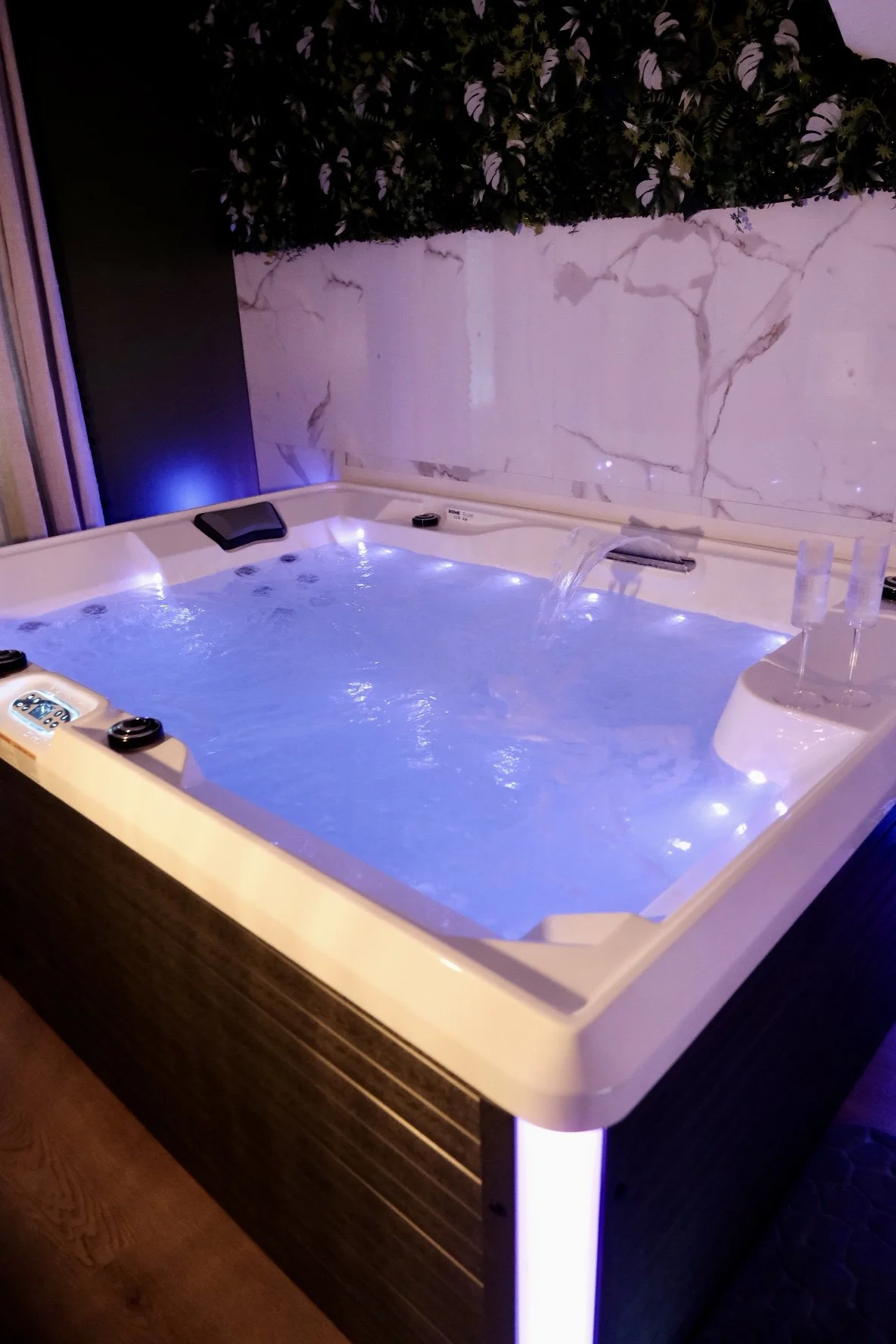 Jacuzzi avec lumières bleues, touches de contrôle, et accessoires comme des verres à champagne, dans une pièce avec murs en marbre et végétation décorative au plafond.