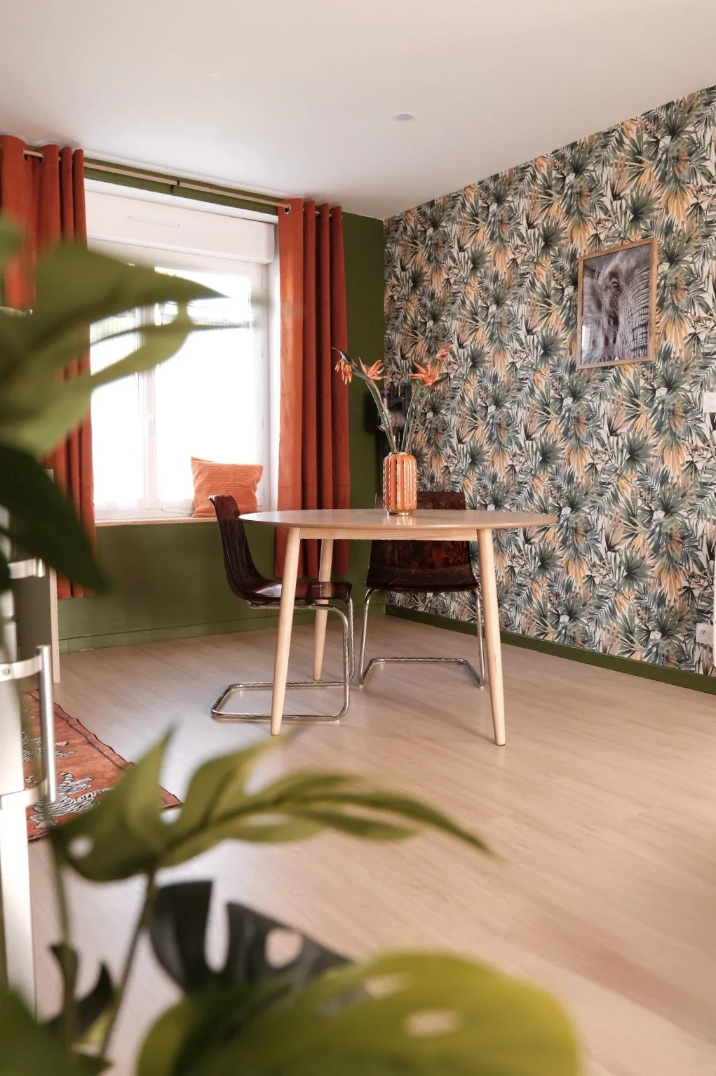 Une salle à manger avec une table en bois, deux chaises en bois foncé, un mur avec un papier peint à motif végétal, une fenêtre avec des rideaux orange et un coussin orange sur le rebord, une peinture d'un éléphant, une bouteille en verre avec des fleurs, un tapis orange, et des murs peints en vert.