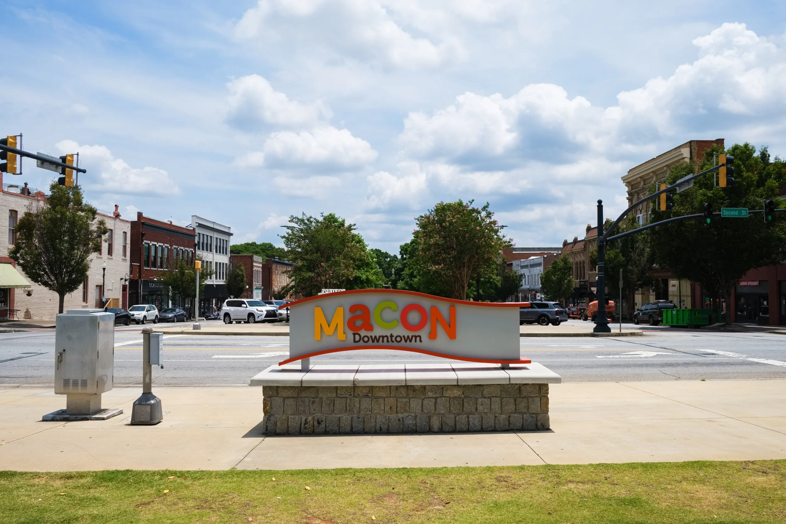 Sign reading 'Macon Downtown' in front of a street with parked cars and buildings, trees, and a partly cloudy sky.