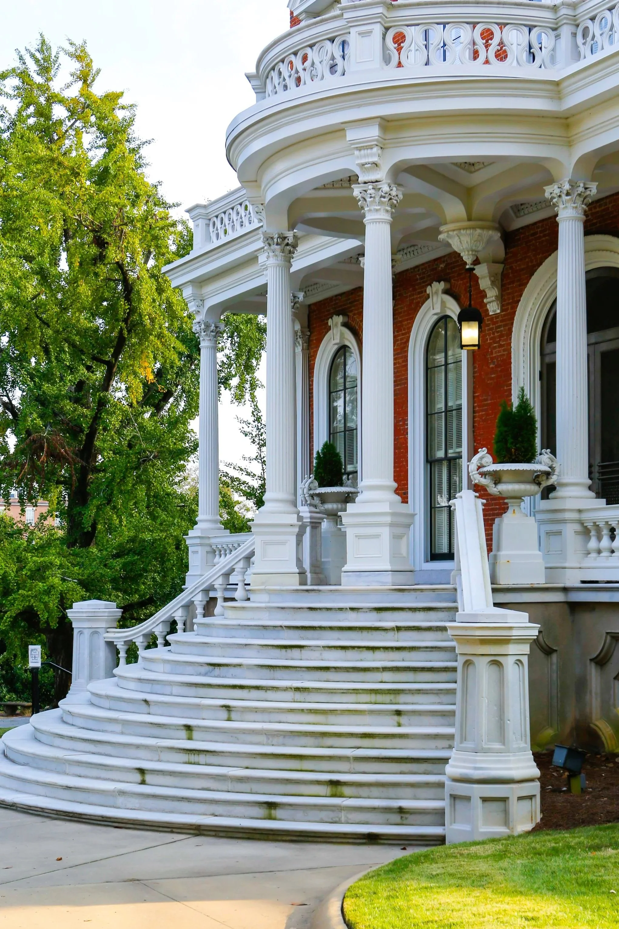 Close-up of the front porch of a historic house with curved marble stairs, tall white columns, and decorative plants in planters, with trees in the background.
