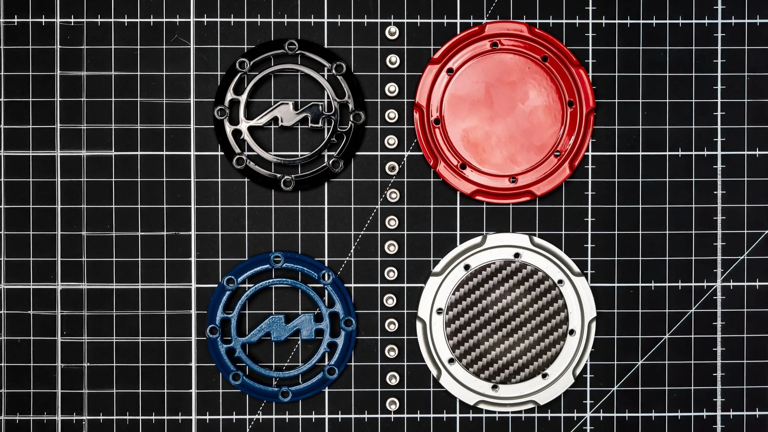 Four circular mechanical parts arranged on a black grid cutting mat: a black part with a lightning bolt and zigzag design, a red part, a blue part, and a metallic silver part with a carbon fiber pattern.