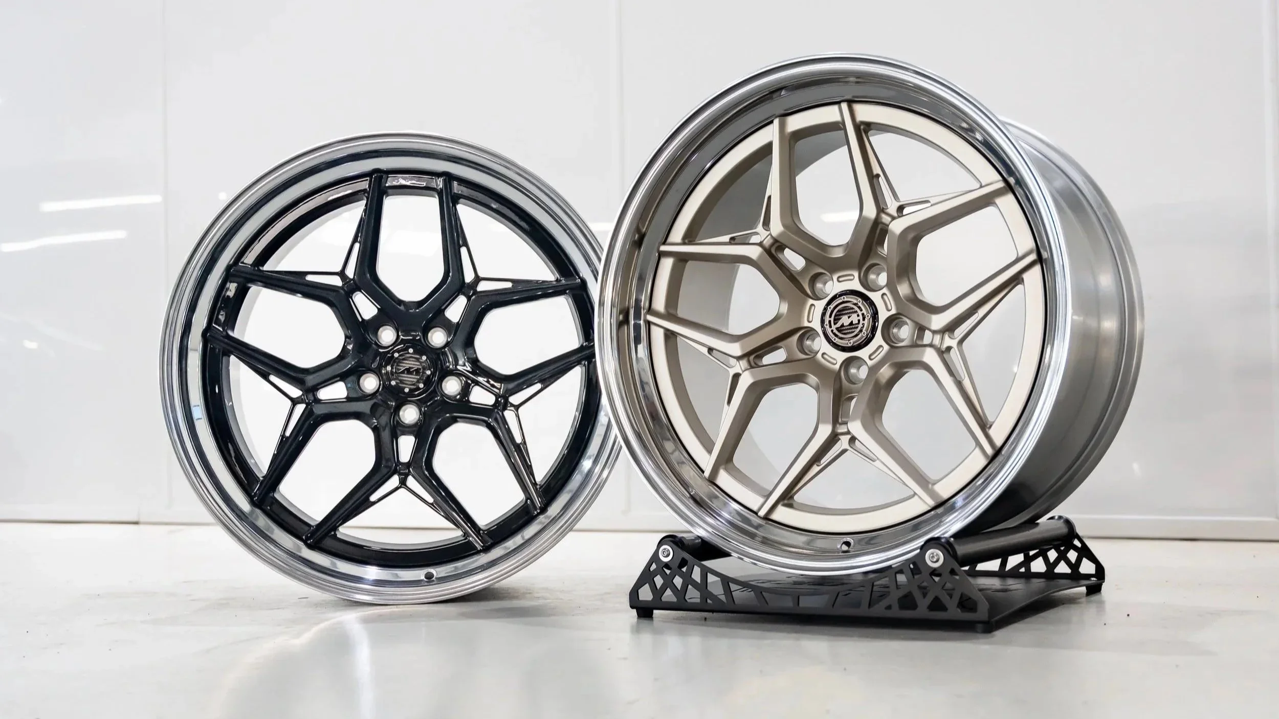 Two automotive wheels with intricate spoke designs, one in black and silver and the other in gold, displayed on a white surface.