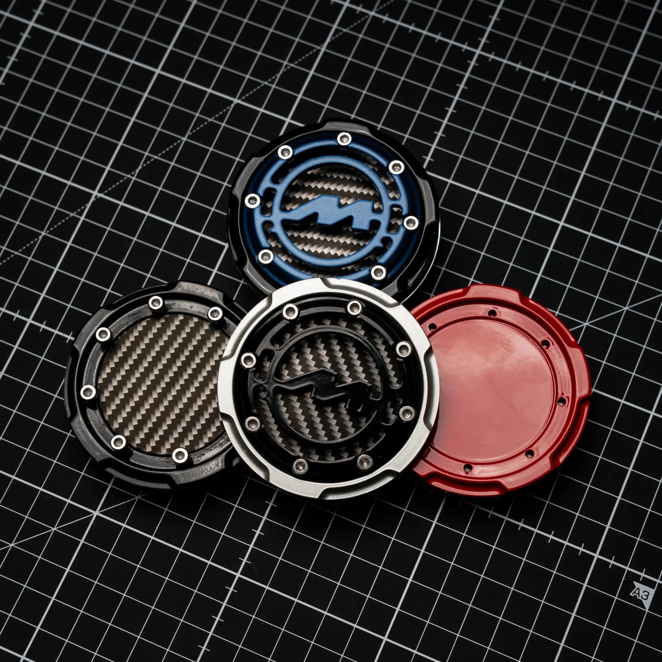 Collection of four colorful watch bezels with black, blue, silver, and red colors, placed on a black cutting mat with white grid lines.