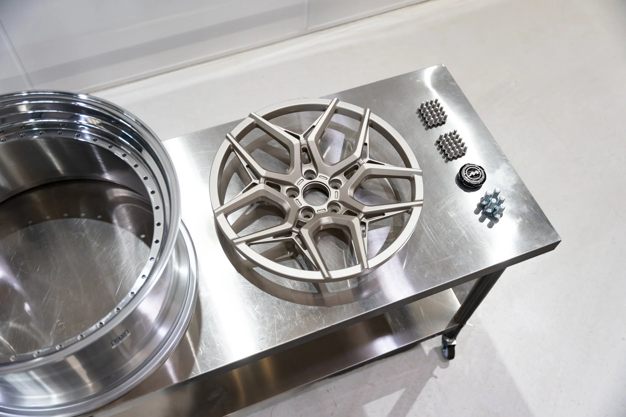 A metal table with disassembled car or bicycle parts, including a large circular alloy wheel, a decorative wheel cover, small metal components, and screws.
