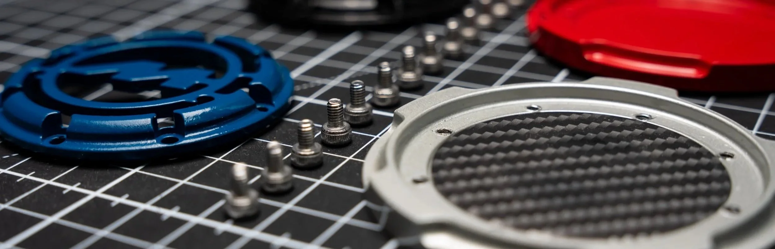 Various mechanical parts including a blue circular component, a white frame with a carbon fiber pattern, red and gray components, and several small screws laid out on a black cutting mat with grid lines.