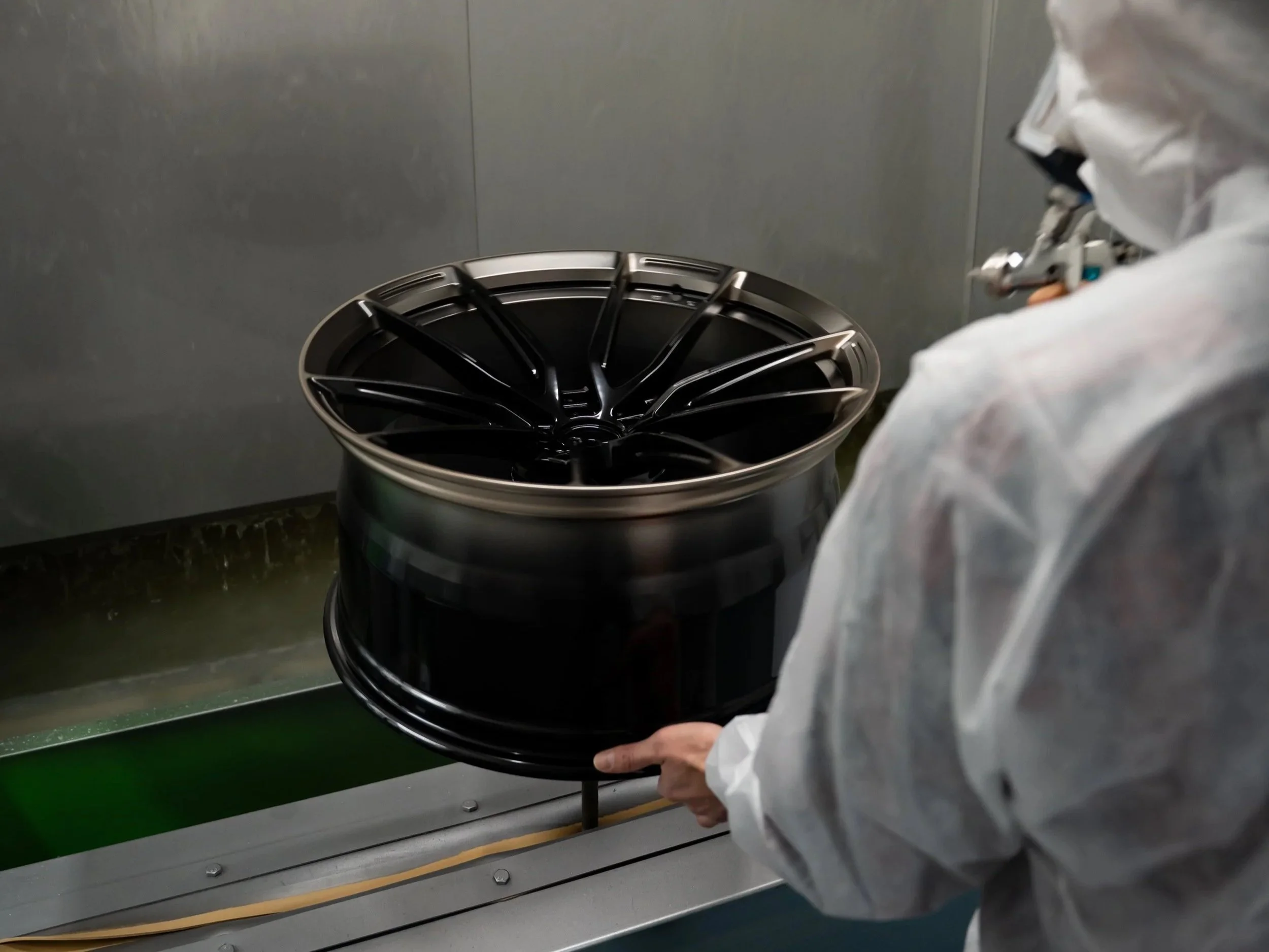 Person in protective clothing handling a black alloy wheel in a manufacturing or painting process.